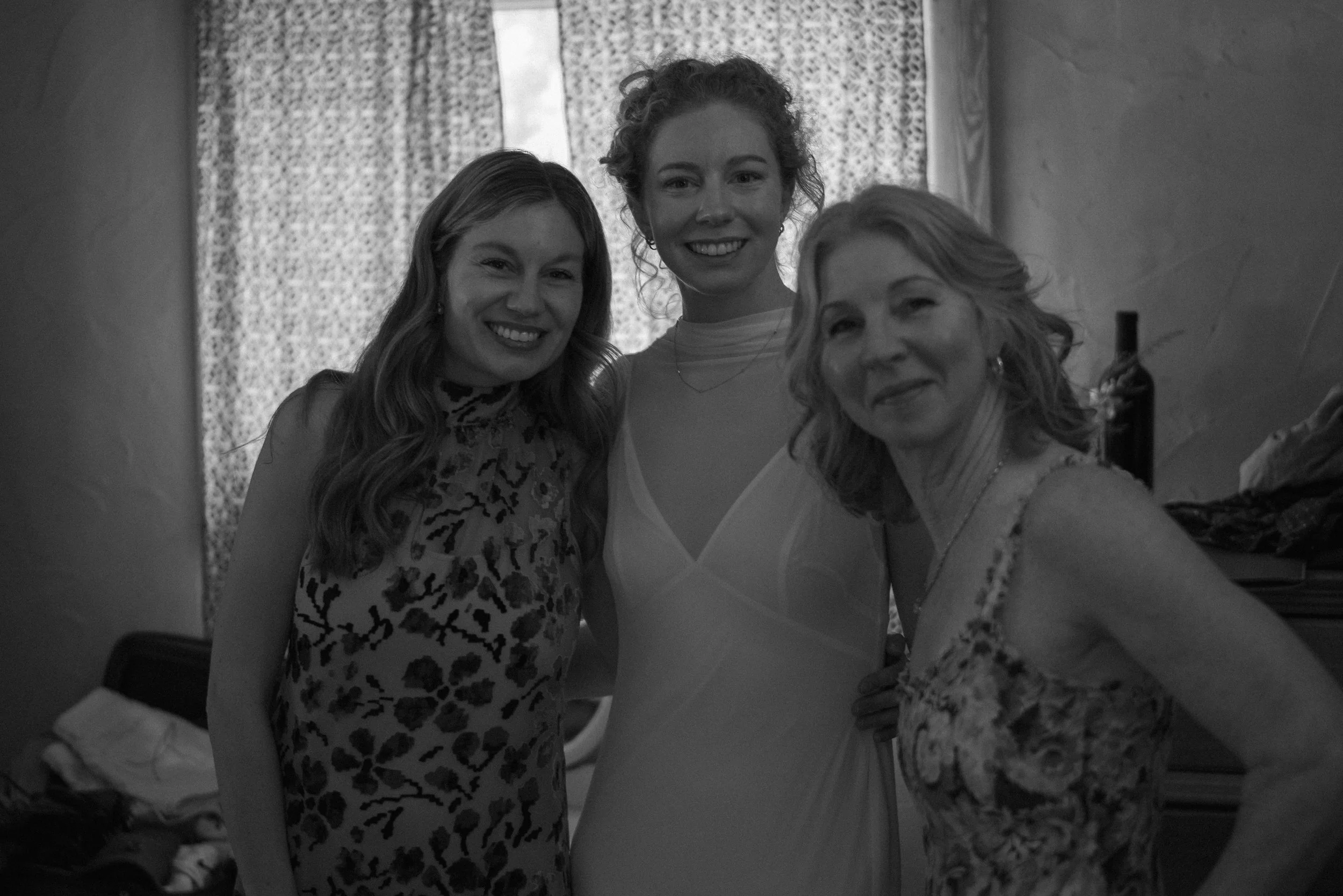 Three women smiling for a photo in front of patterned curtains. They are dressed nicely, with one in a leopard print top, one in a light-colored dress, and the third in a floral patterned top.