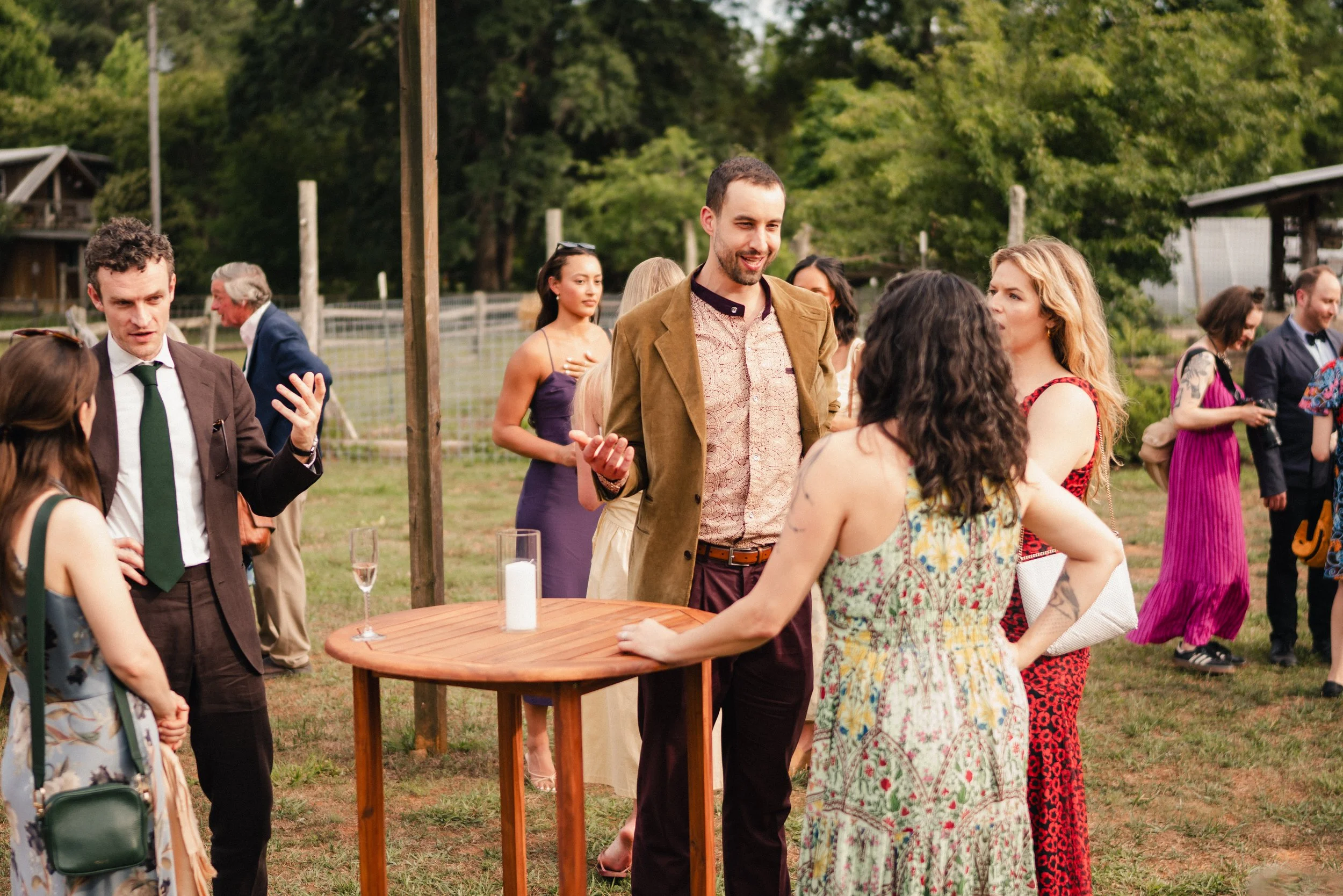 Group of people socializing outdoors at a garden party, with green trees in the background and standing around a wooden table.