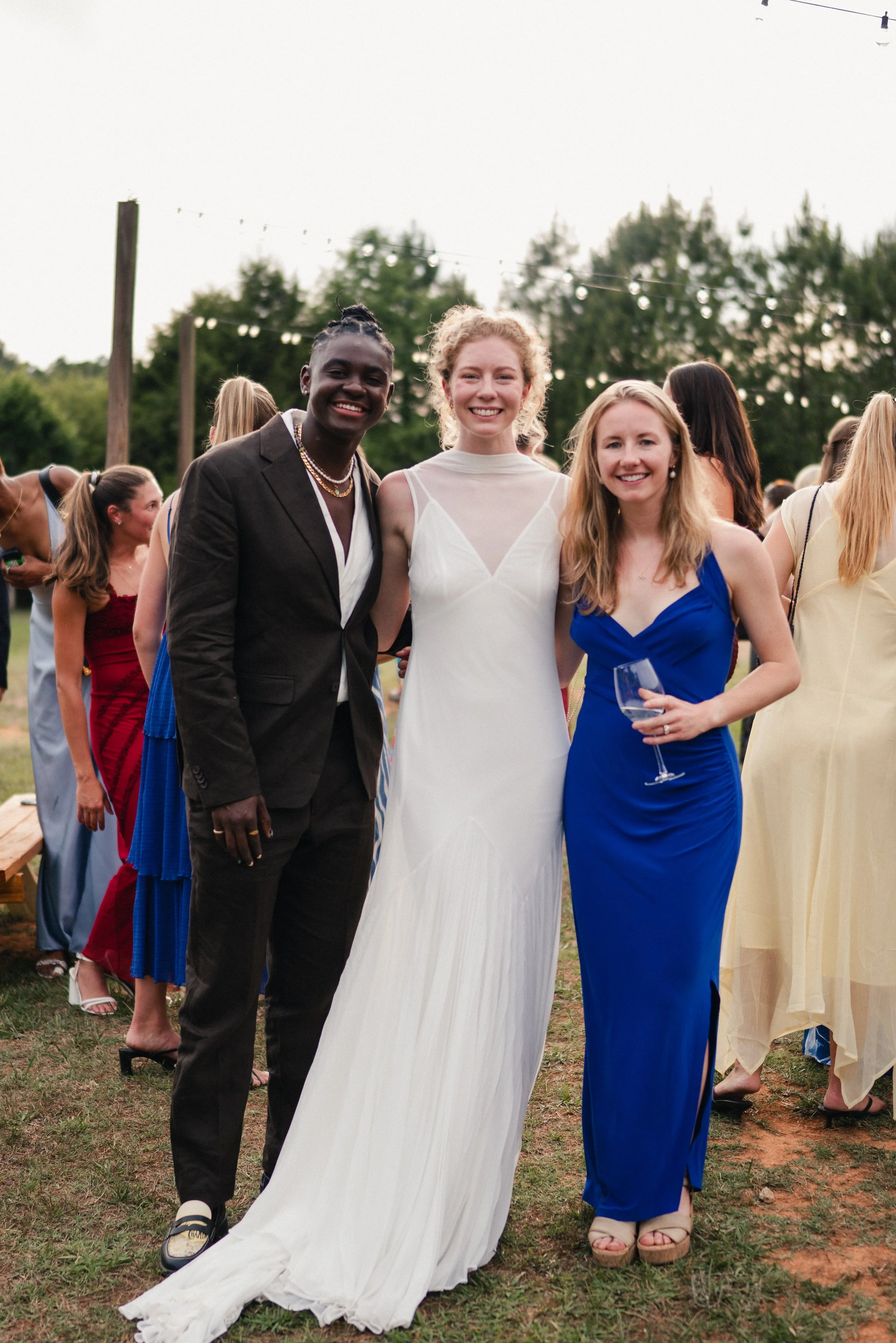 A group of diverse women at an outdoor wedding or celebration, with three women in the foreground dressed in elegant evening attire, smiling and holding glasses, with other guests in the background and string lights overhead.