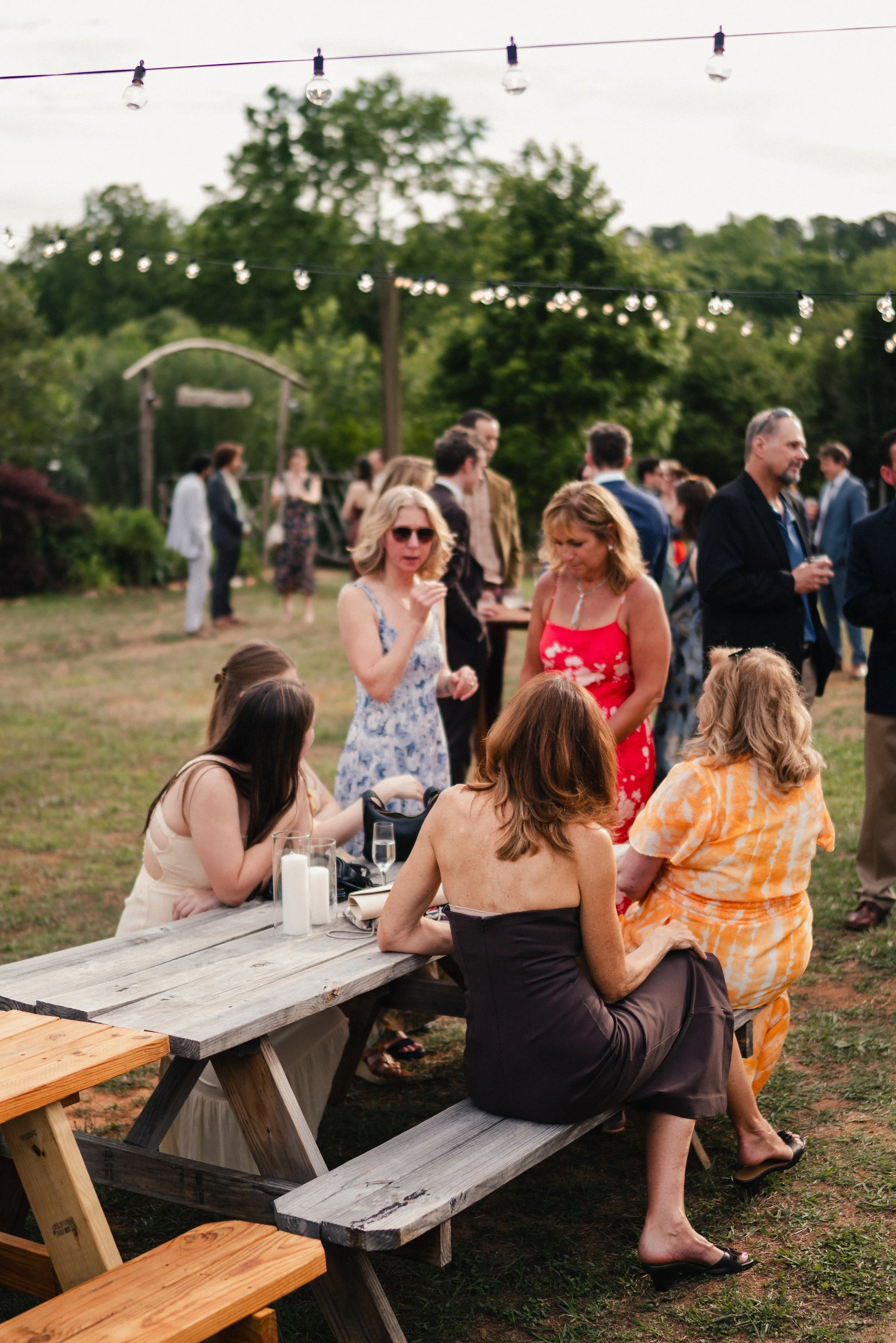 People at an outdoor gathering in the evening, some sitting at a wooden picnic table with glasses and candles, others standing and talking under string lights, with trees and a cloudy sky in the background.