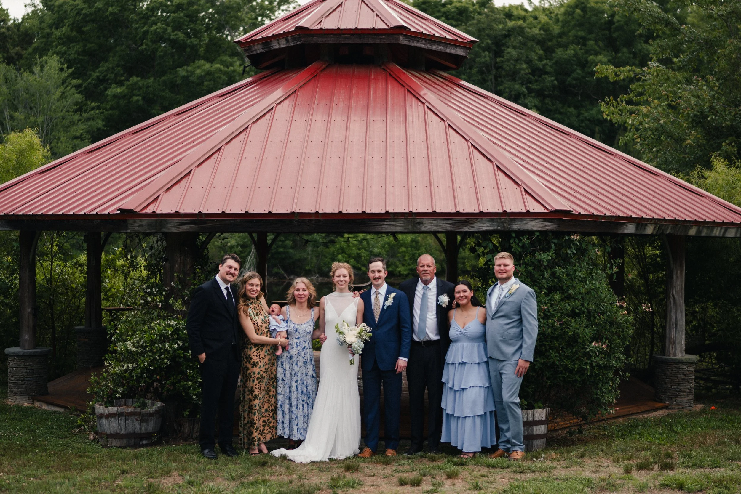 A group of people, including a bride and groom, posing outdoors in front of a gazebo with a red metal roof during a wedding celebration.