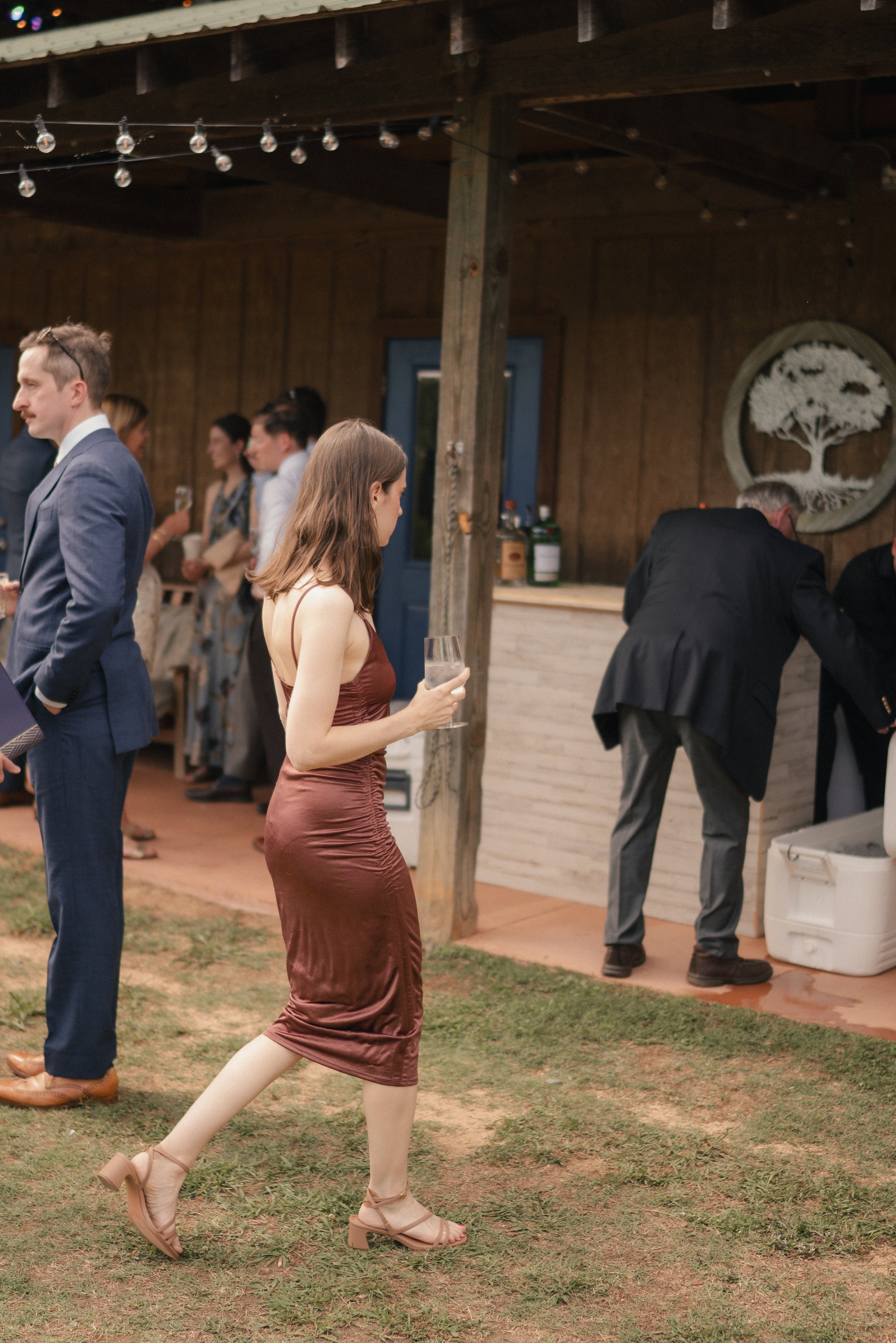 A woman in a brown satin dress and beige high-heeled shoes walking on a grassy area at an outdoor event, with other people in formal attire in the background near a wooden building with string lights and a decorative tree artwork on the wall.