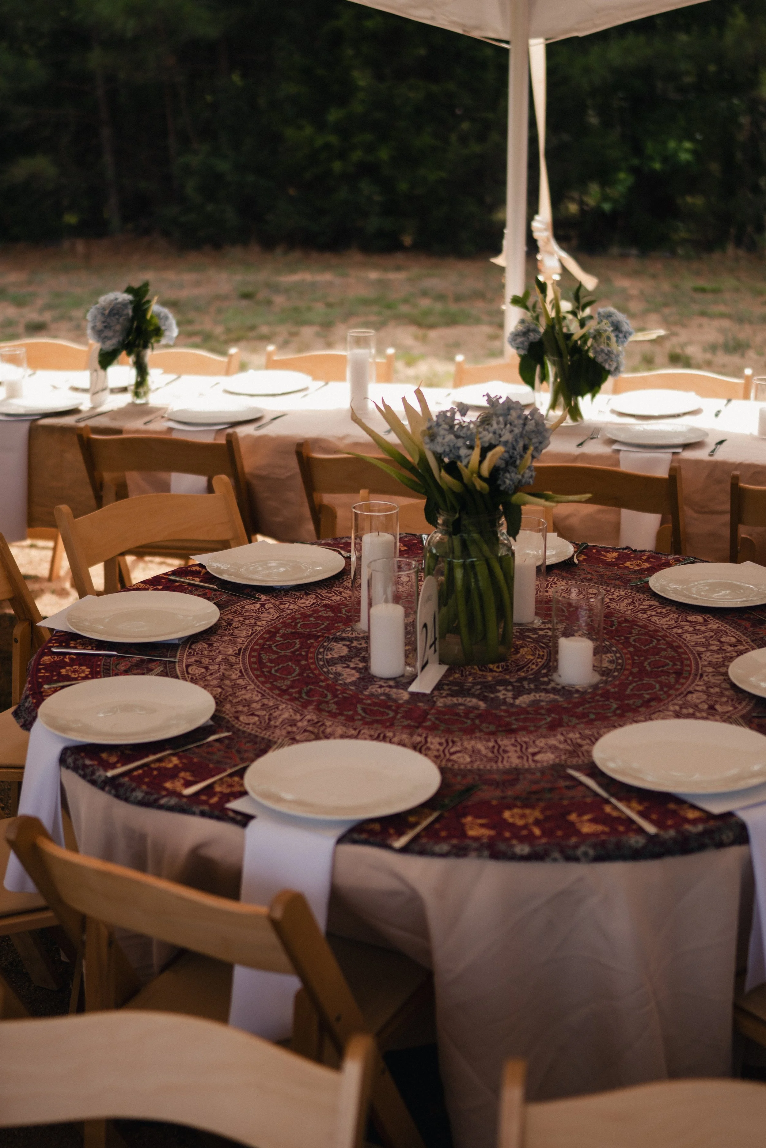 An outdoor dining table set for a meal with plates, silverware, wine glasses, and floral centerpieces under a large white canopy, with a forest in the background.
