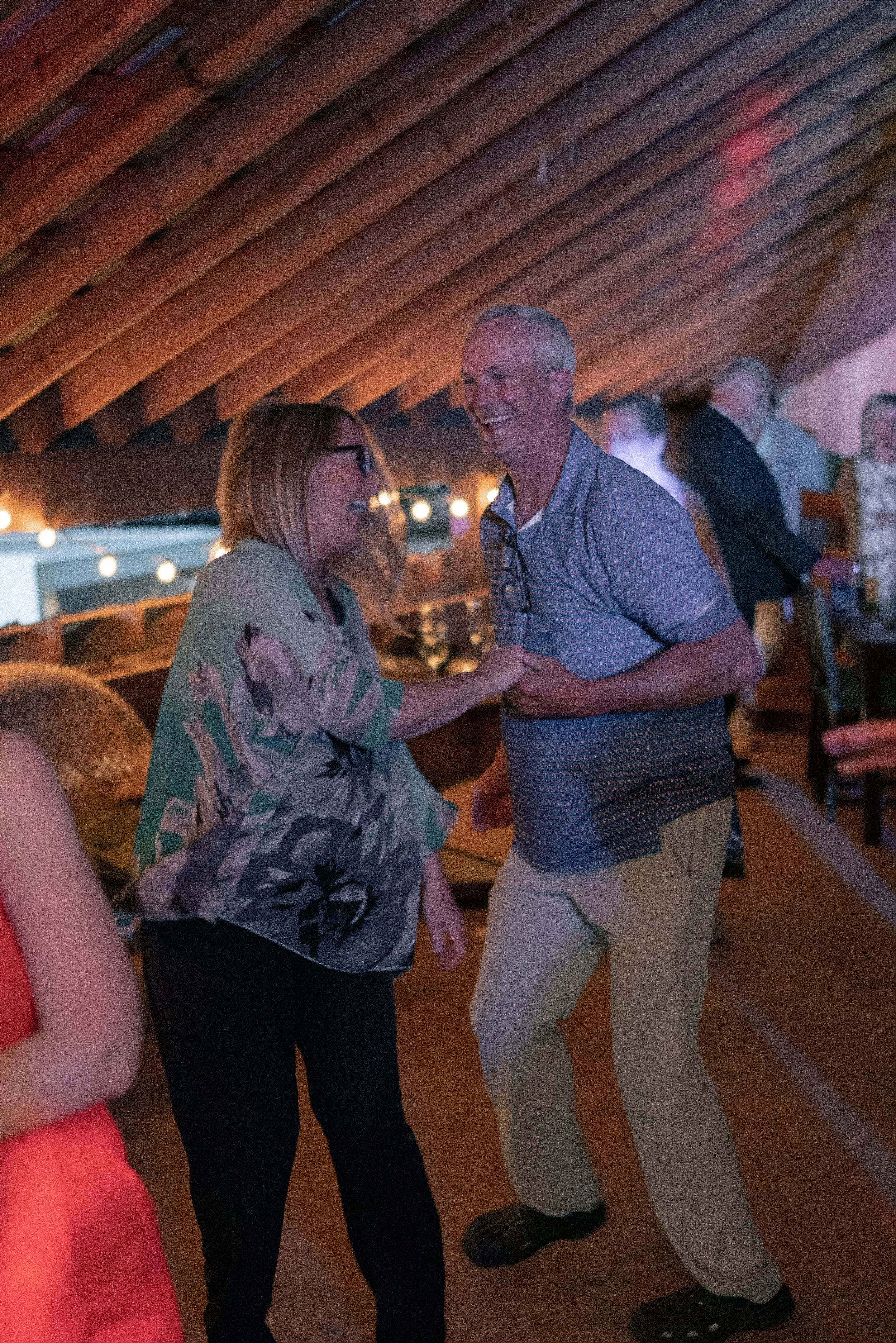Two middle-aged adults, a woman with long blonde hair and glasses, and a man with short white hair, dancing and laughing in a warmly lit indoor setting. Other people are in the background, and the ceiling is made of wooden beams.
