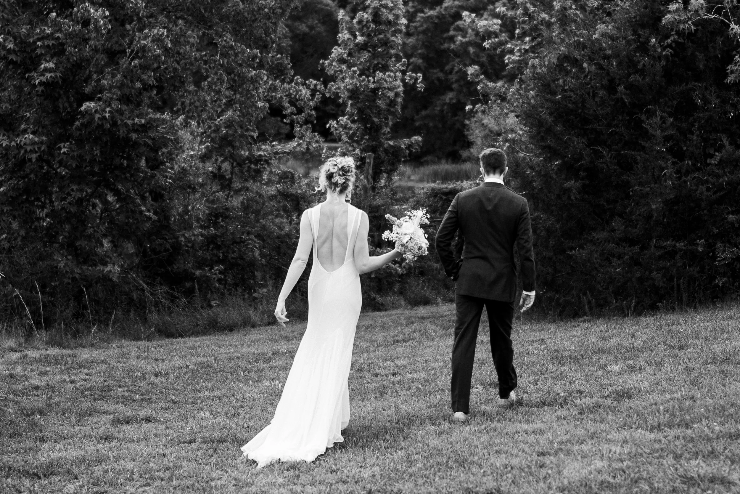 A wedding couple walking away through a field, the bride in a long wedding dress holding a bouquet, the groom in a suit, with trees in the background.
