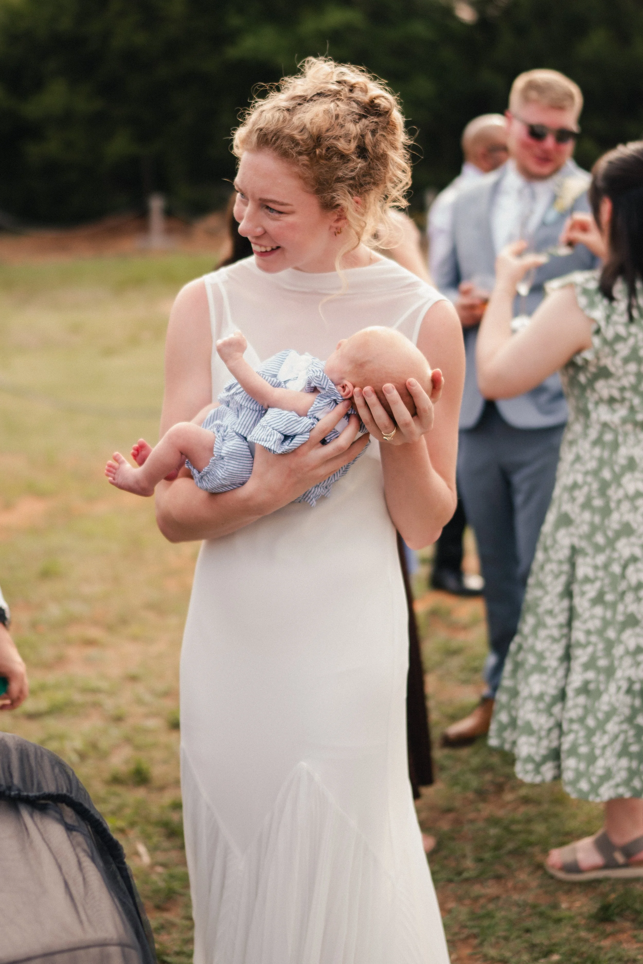 A woman with curly red hair wearing a white dress, holding a baby in a blue striped outfit, at an outdoor gathering with people in the background.