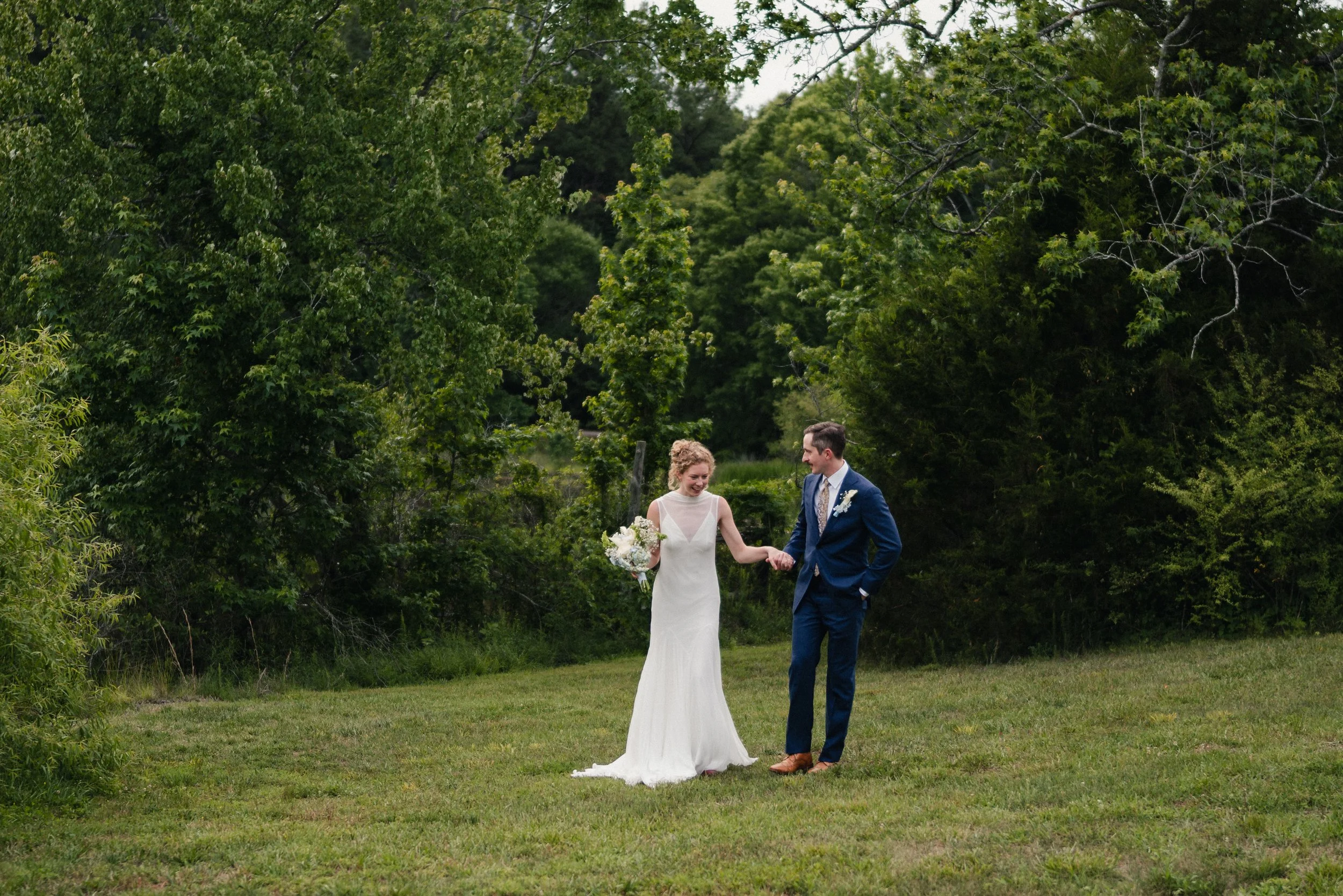 A bride and groom holding hands and walking through a grassy outdoor area surrounded by green trees, on their wedding day.