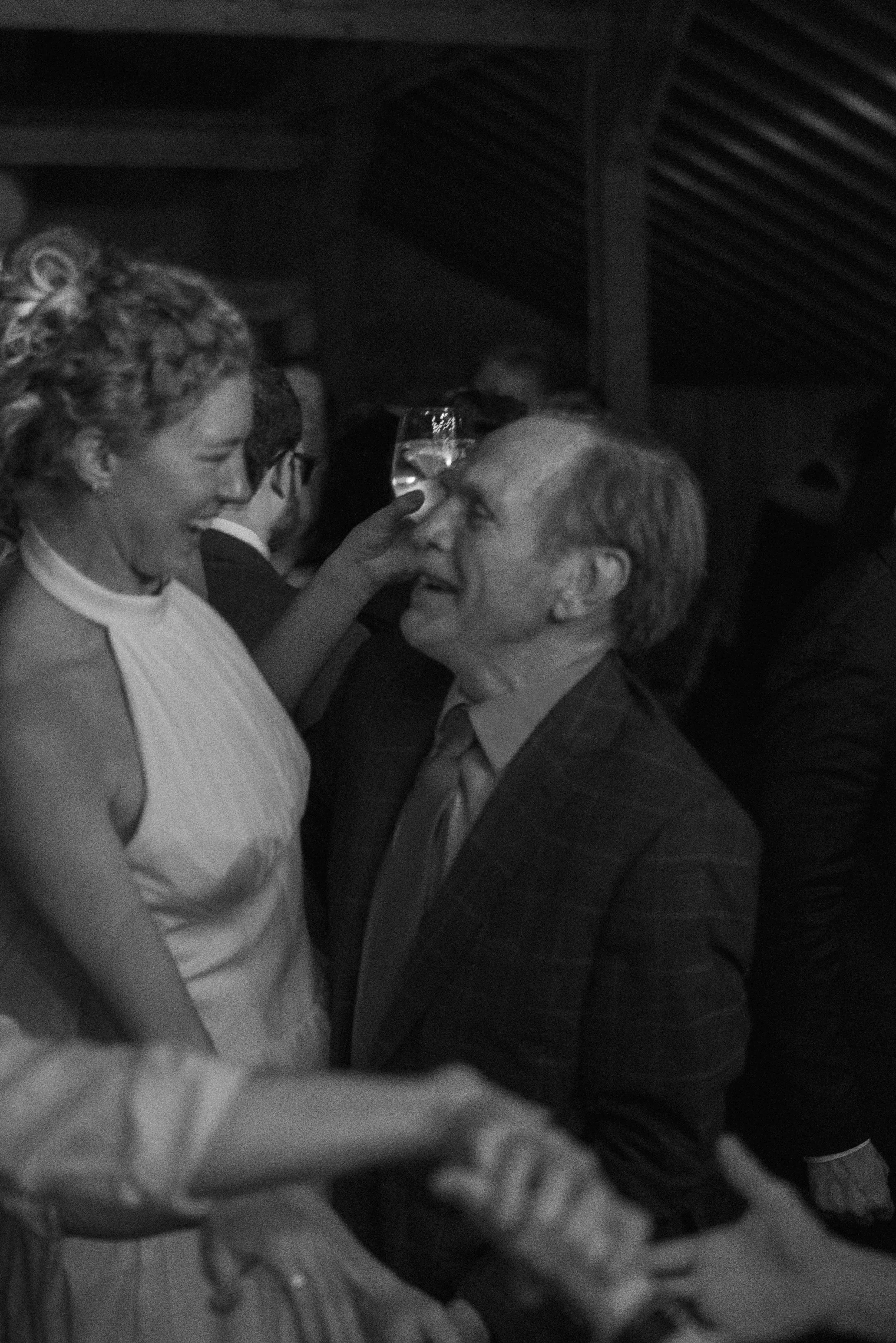 A woman with curly hair and a light-colored sleeveless top is smiling and touching foreheads with a man who has short hair and is wearing a checked blazer and dress shirt. They are in a dark indoor setting with some light reflections behind them.