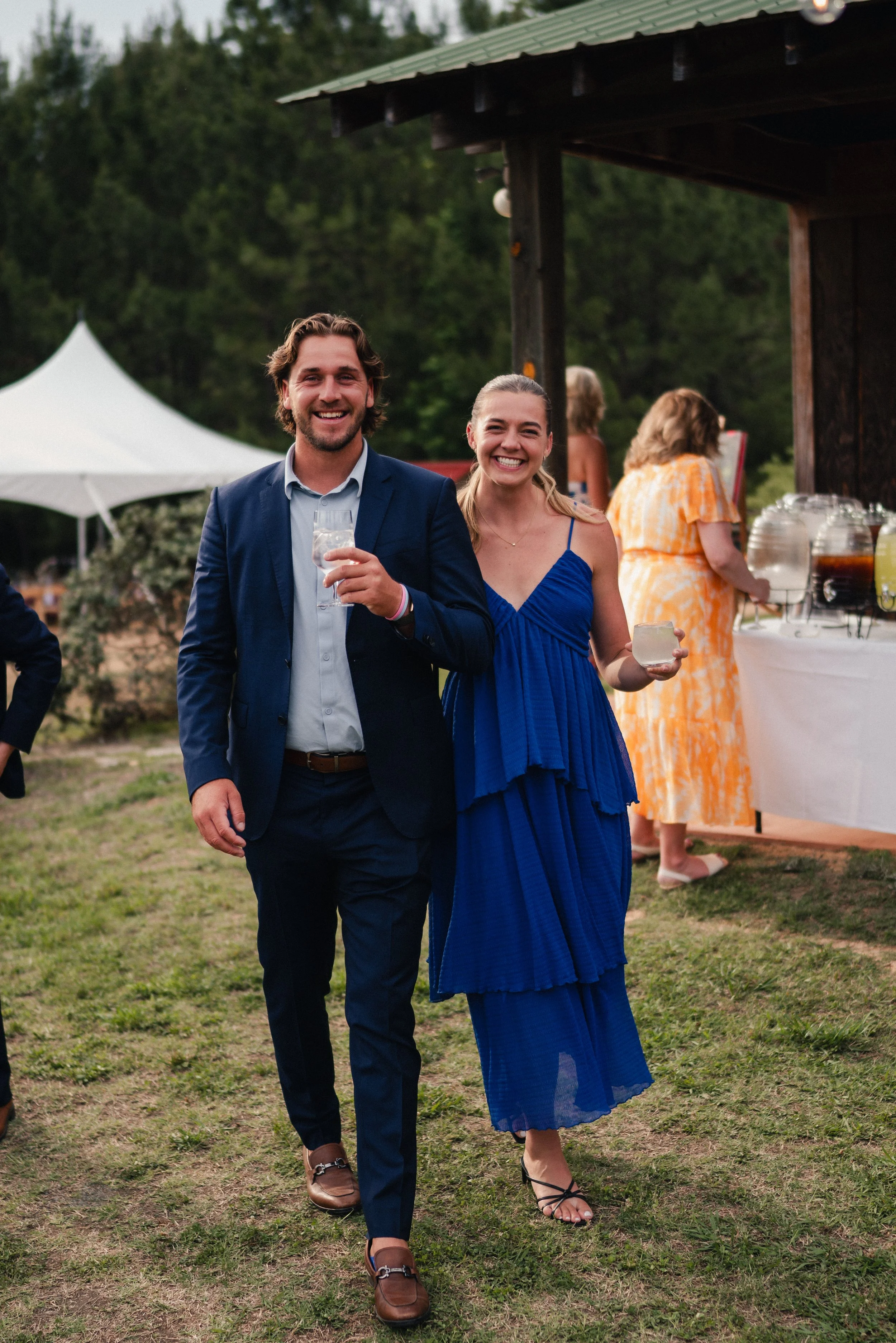 A man and woman dressed in formal attire walking on grass at an outdoor event, smiling and holding drinks, with a buffet table and people in the background.