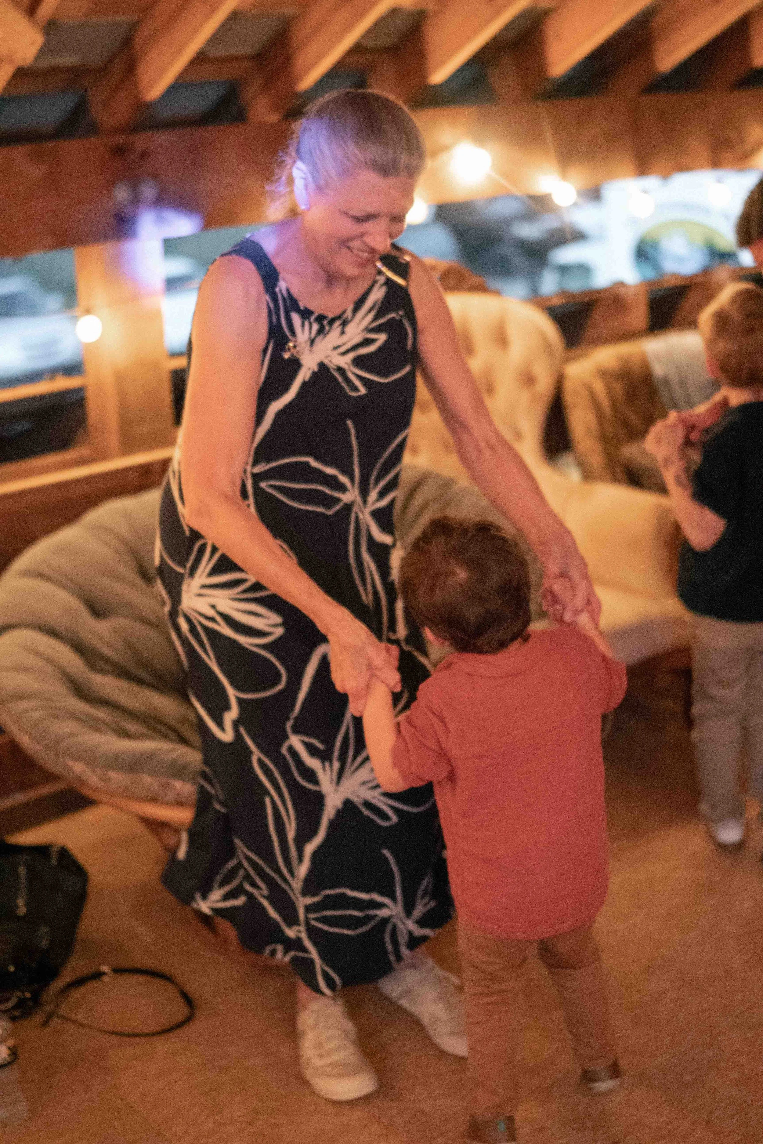 Woman with a floral dress holding hands with two children in a cozy indoor setting with warm lighting and wooden beams.