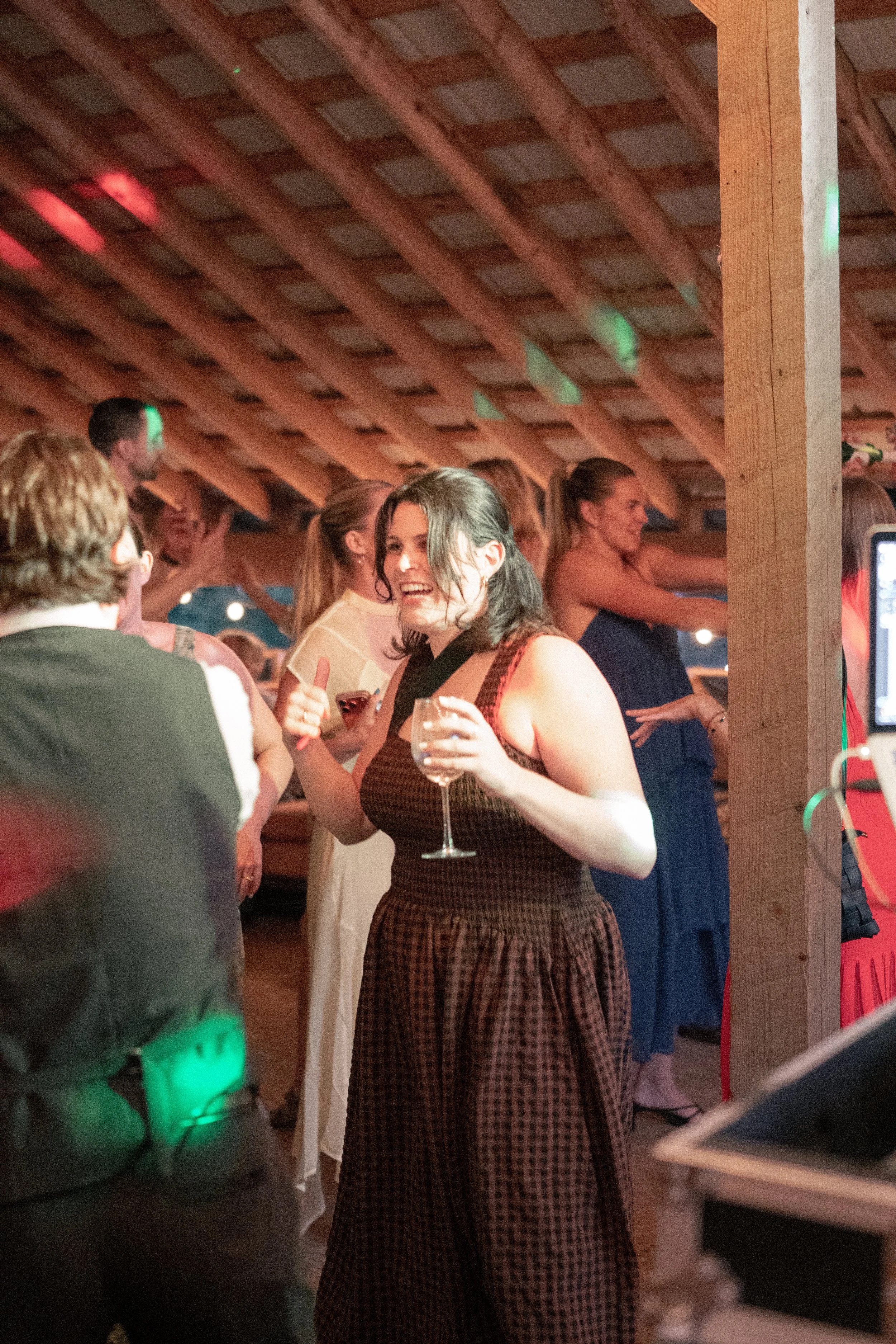 People socializing at a party inside a wooden venue, some holding drinks, with a cheerful woman in the center smiling and giving a thumbs-up gesture.