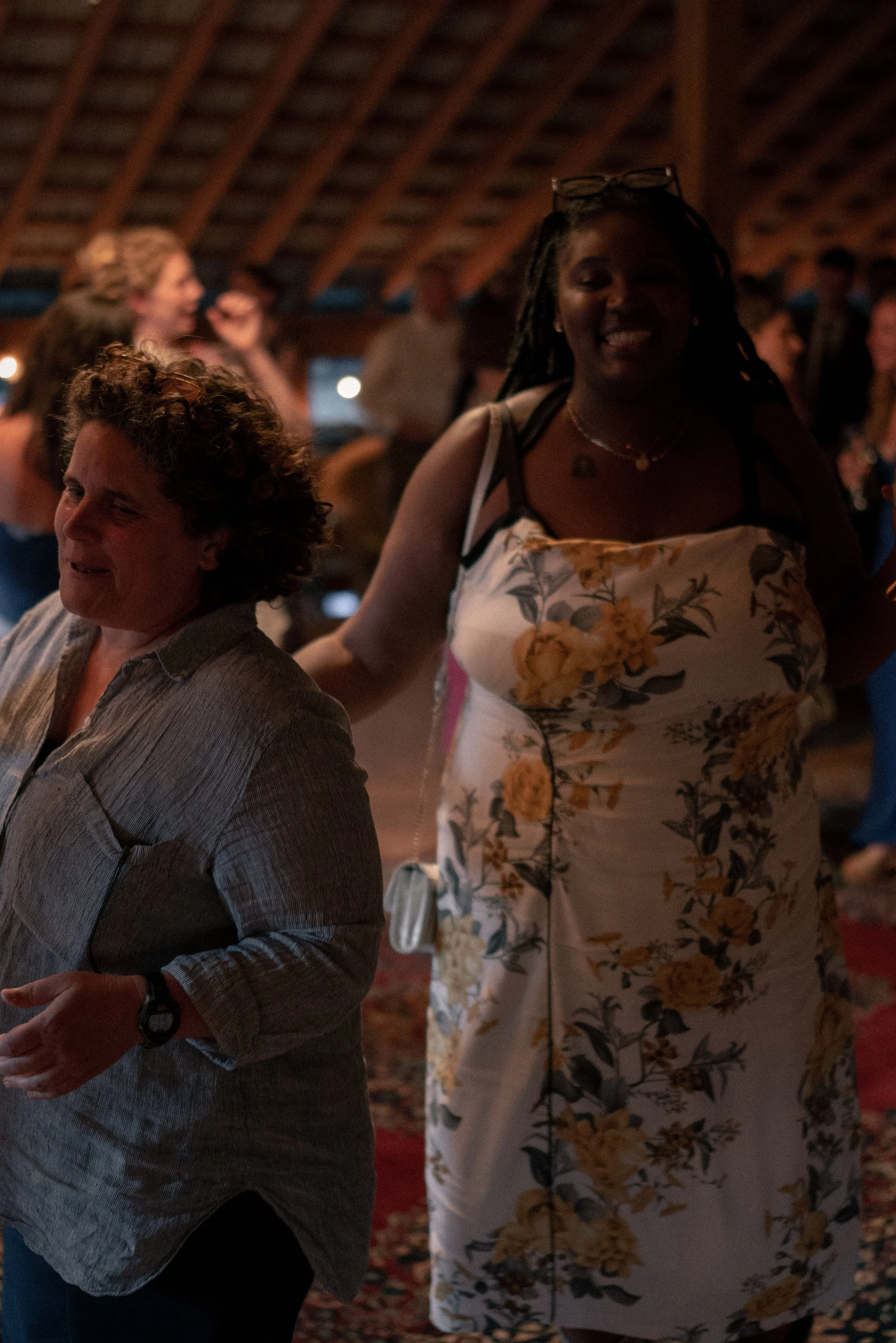 People gathered indoors at a social event, with a woman in the foreground wearing a floral dress smiling, and a man nearby in a gray shirt, with several others in the background.