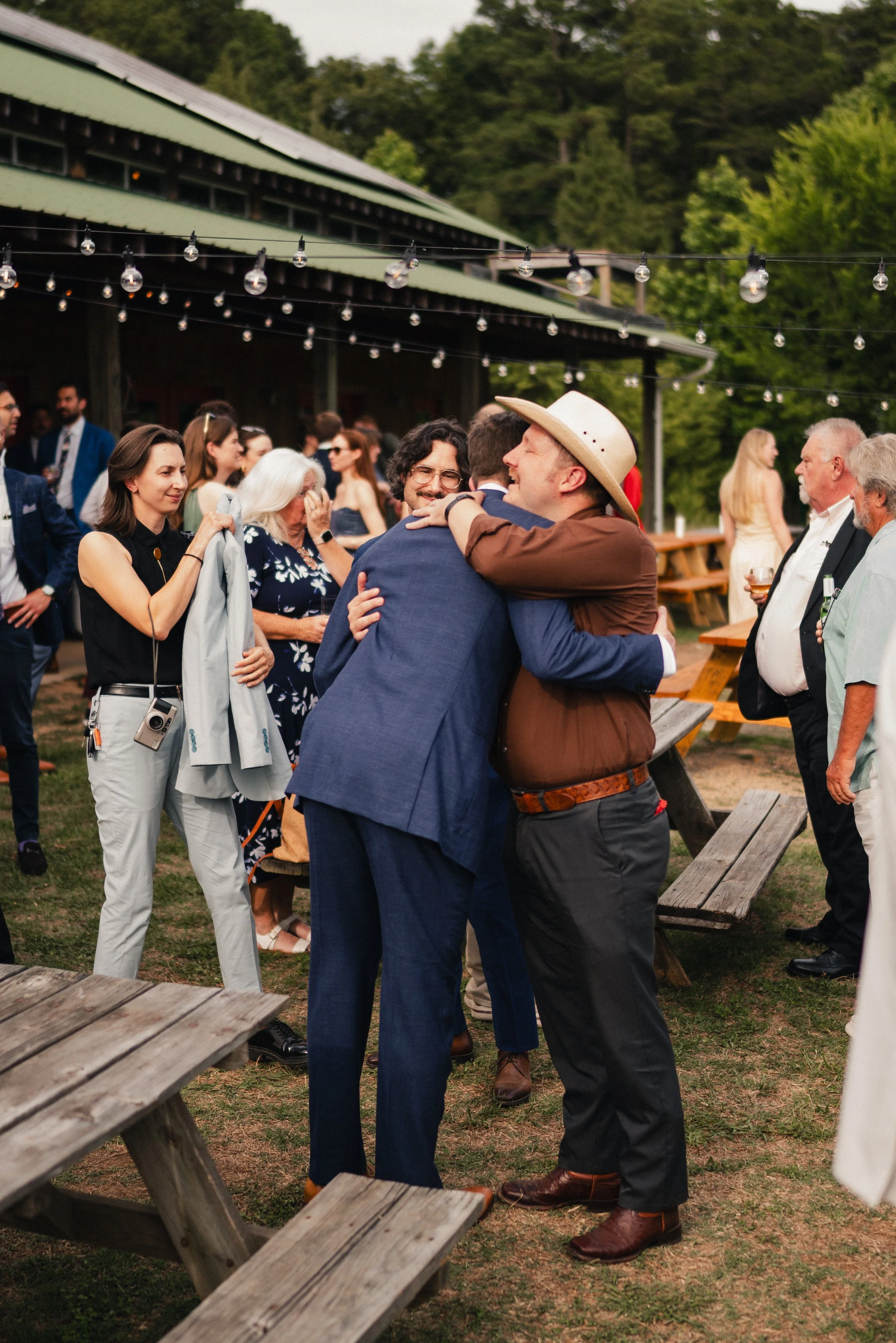 People at an outdoor event, some hugging and chatting among wooden picnic tables, with string lights overhead and green trees in the background.
