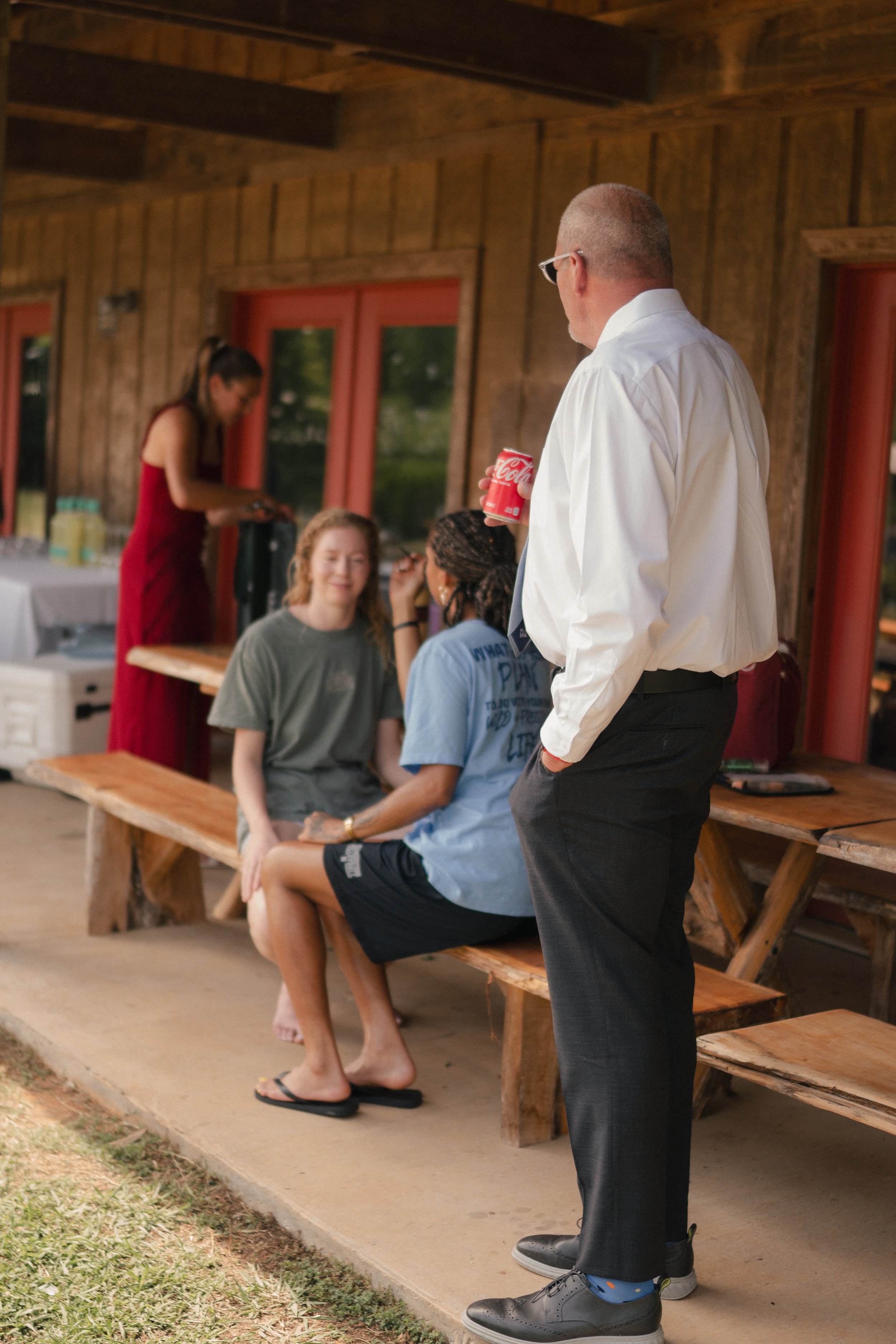 A man in formal attire holding a Coca-Cola can stands near three women sitting on a wooden bench outside a building with wooden walls and red window frames. One woman is getting her makeup done, and the other two are smiling.