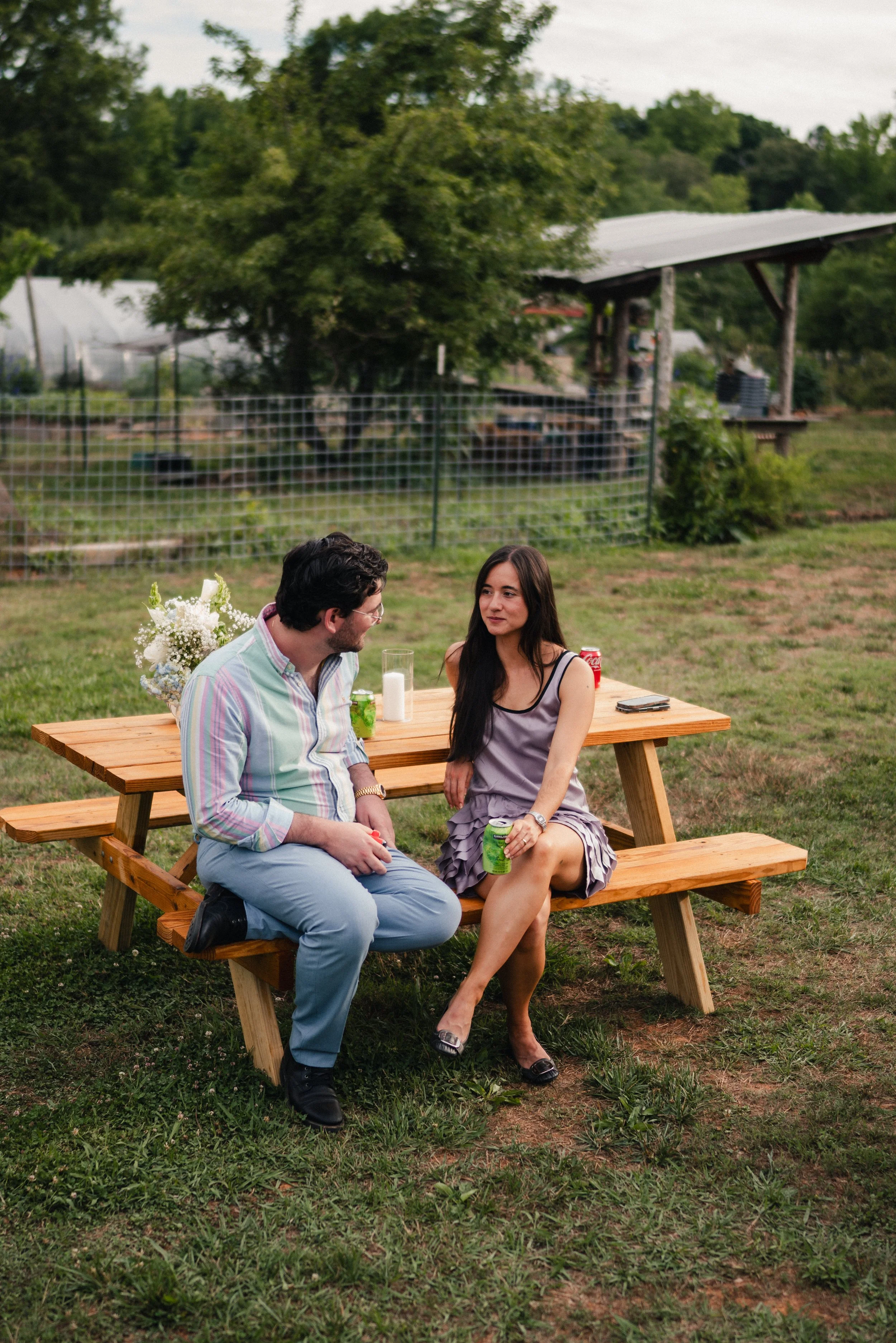 A man and woman sit on a wooden picnic table in a grassy outdoor area, engaging in conversation, with drinks in their hands. Behind them, there is a chain-link fence, trees, a shed with a metal roof, and a partly cloudy sky.