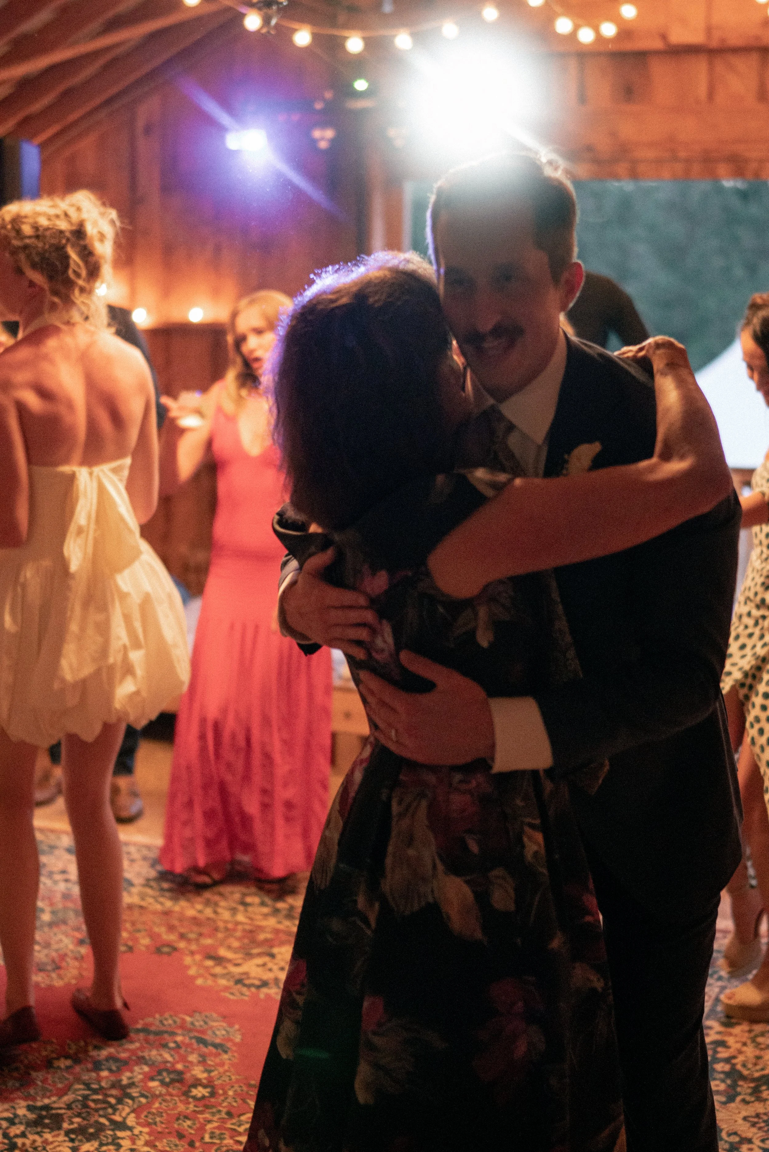 People dancing and socializing in a warmly lit wooden venue, with a man and woman in the foreground hugging, and others in the background enjoying the event.
