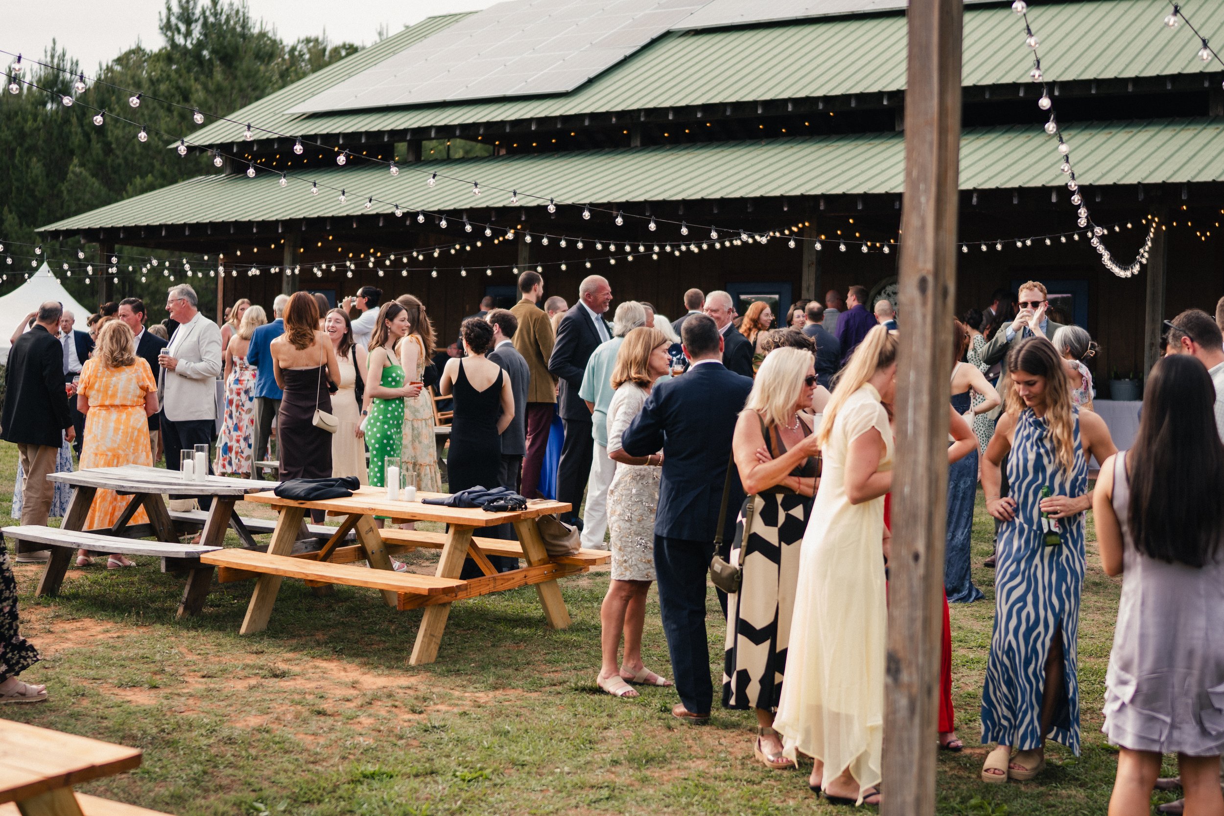Crowd of people socializing at an outdoor event under string lights near a green metal-roofed building.