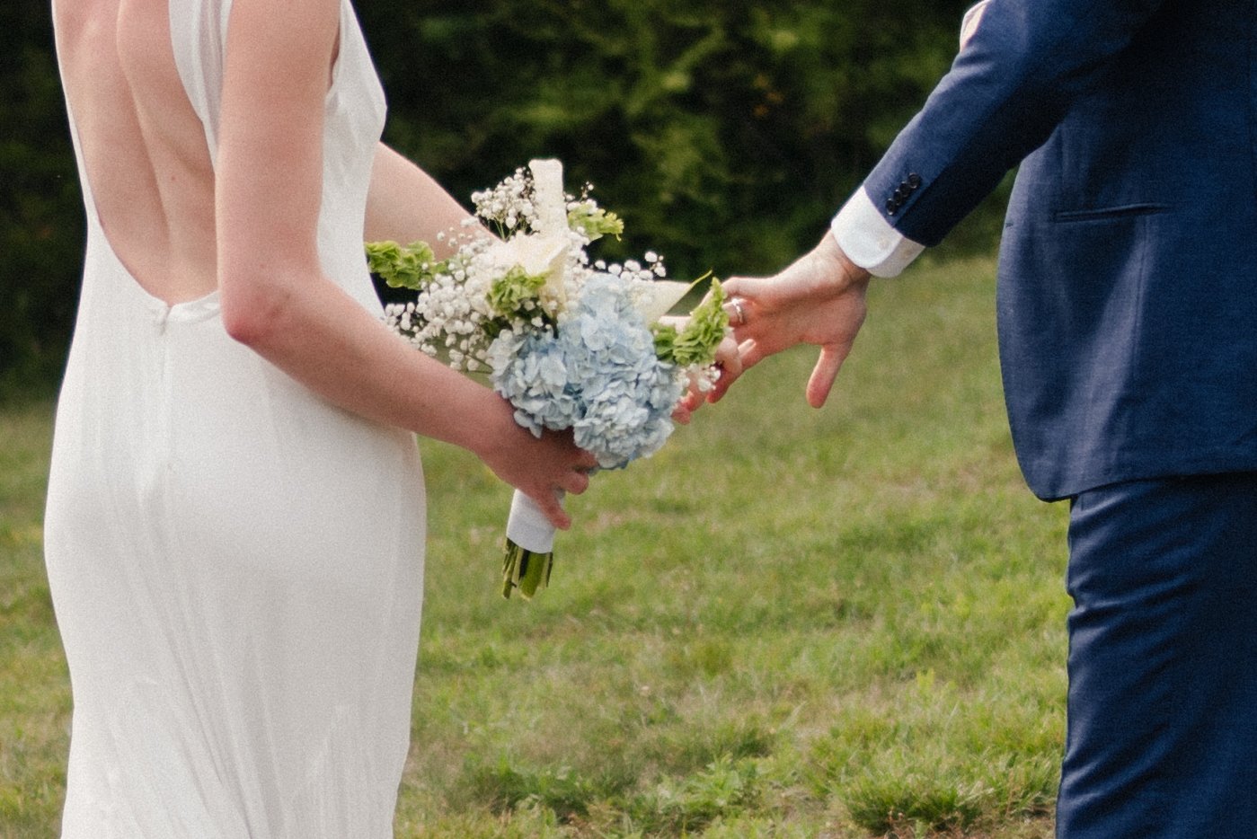 A bride in a white wedding dress holding a bouquet of flowers as a groom in a blue suit extends his hand towards her during an outdoor wedding ceremony.
