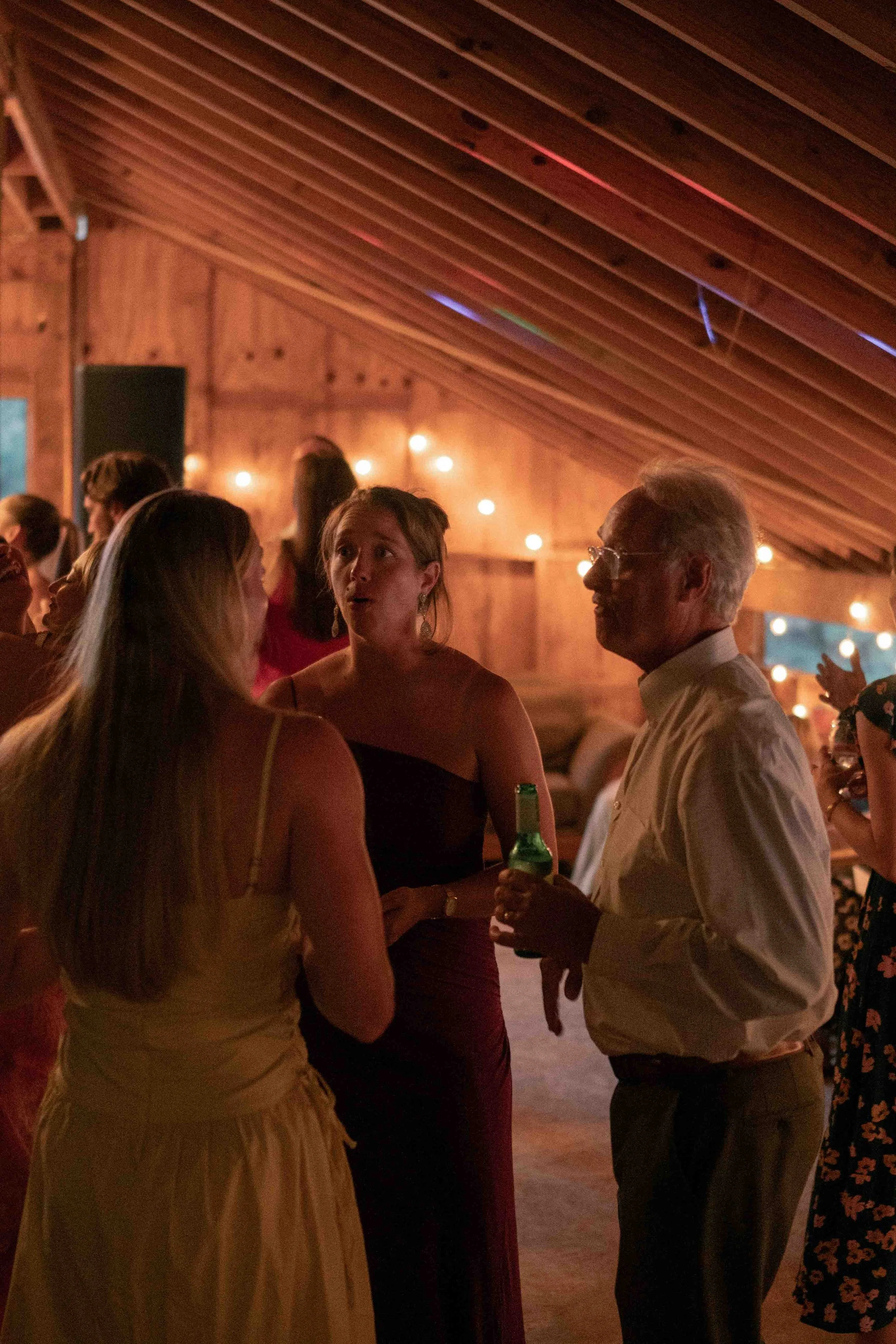 People socializing at an indoor party with warm lighting and wooden walls, dressed in semi-formal attire, engaged in conversation.
