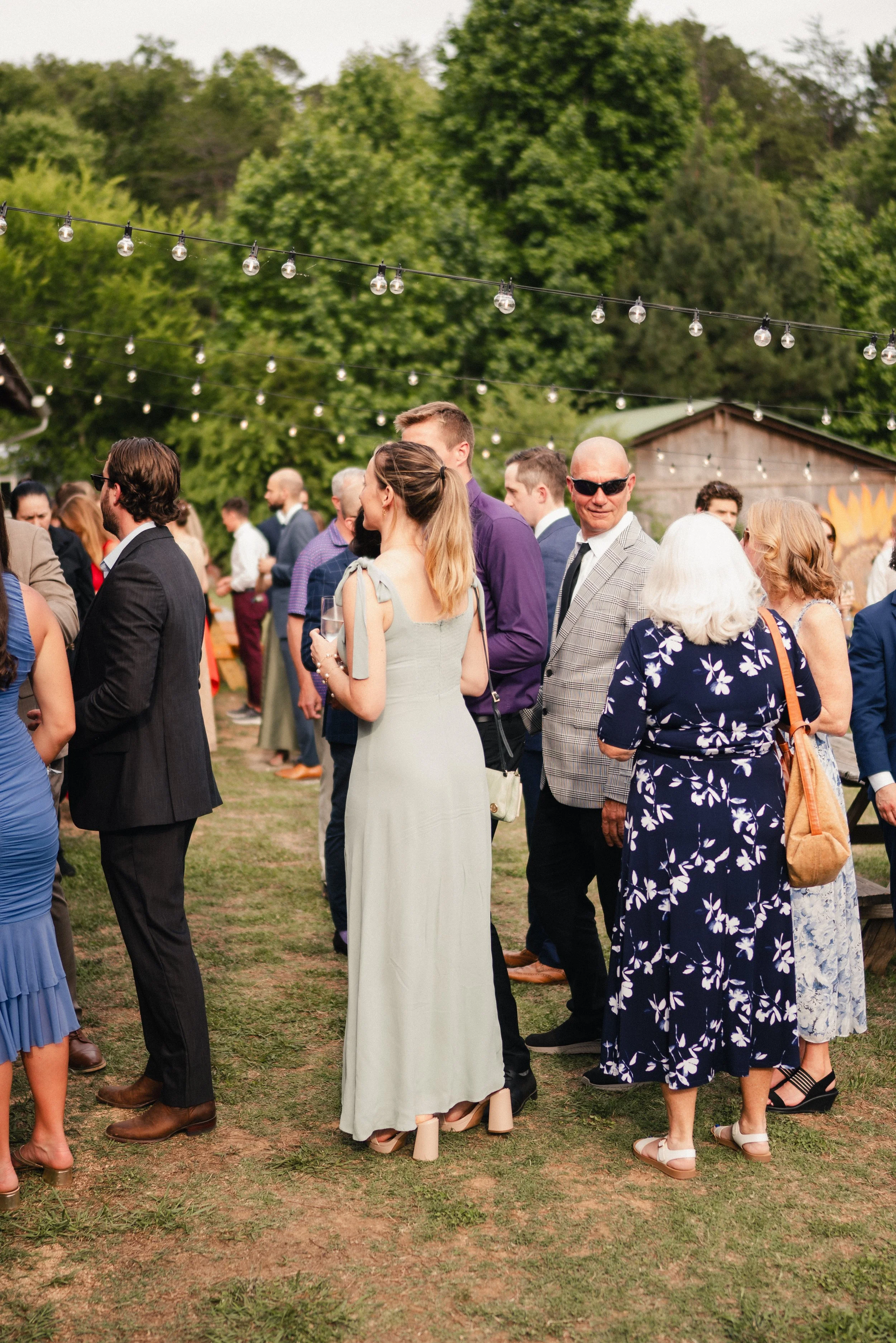 People gathered outdoors at a social event, dressed in semi-formal attire, with string lights hanging above and green trees in the background.