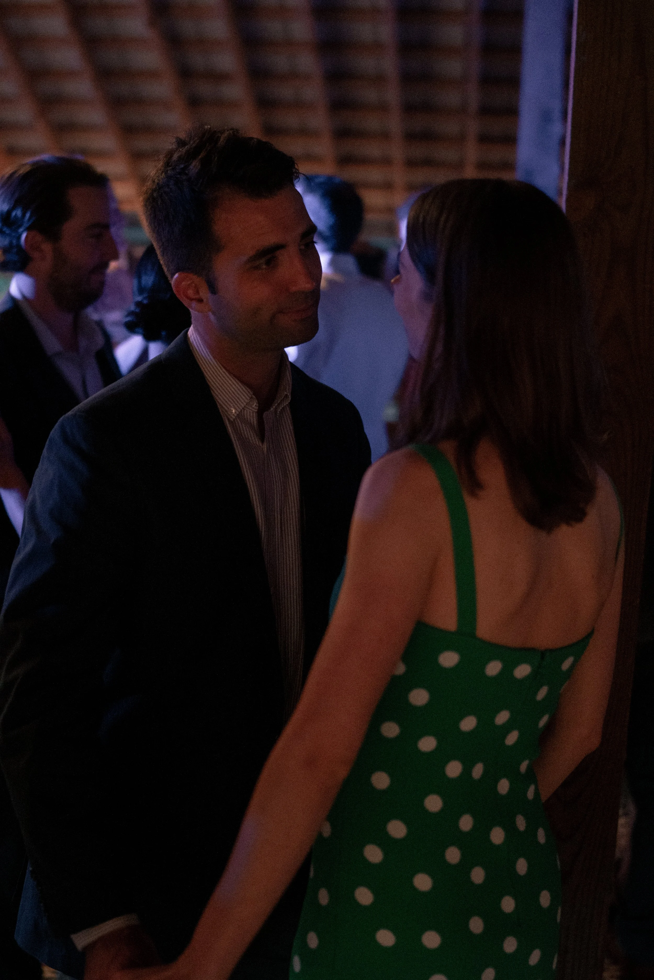A man and a woman are standing close to each other and talking. The man is wearing a suit jacket and a collared shirt. The woman is wearing a green dress with white polka dots. In the background, there are other people and a wooden wall.