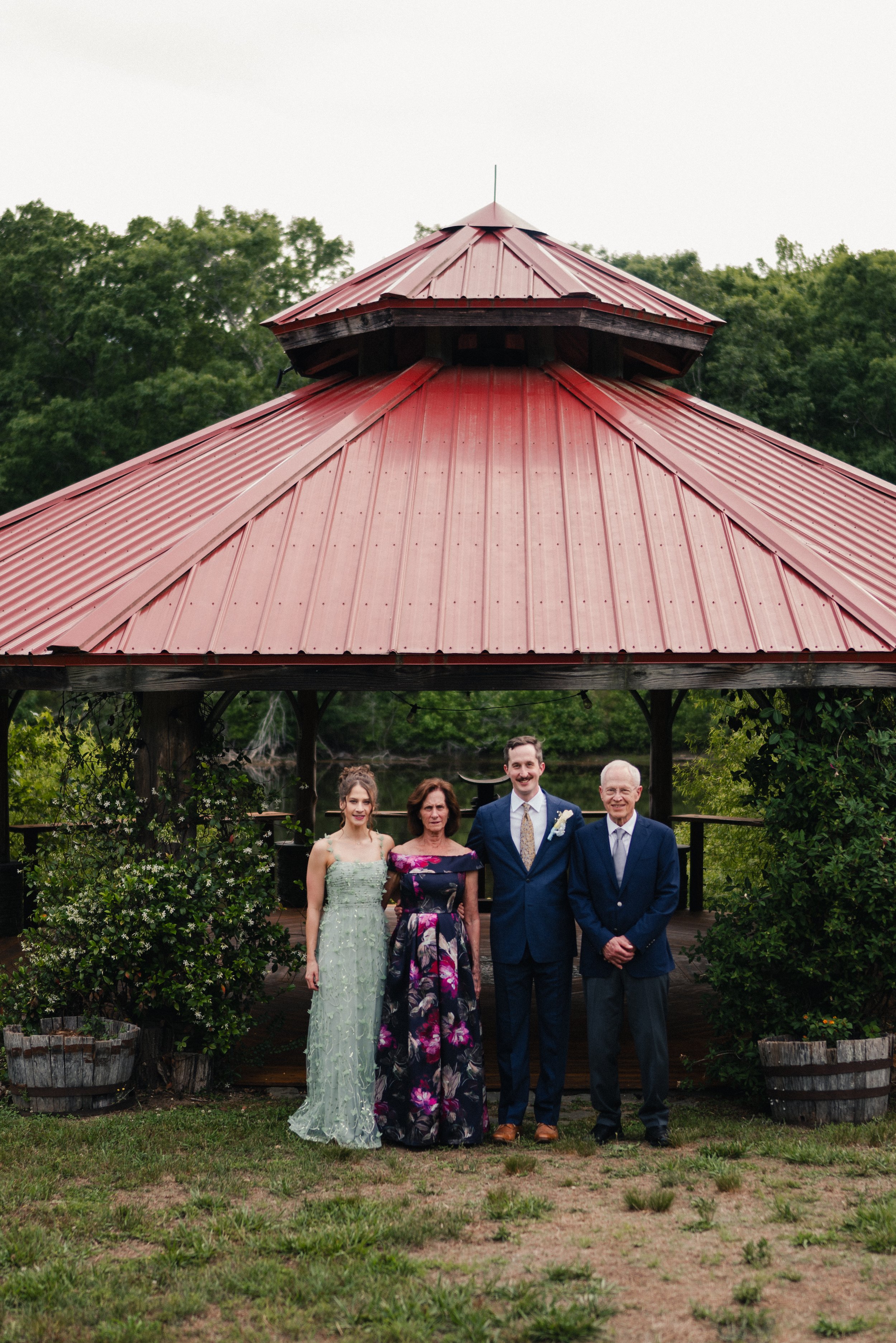 A group of five people, three women and two men, standing under a red metal gazebo in a garden setting with trees and greenery, dressed in formal attire.