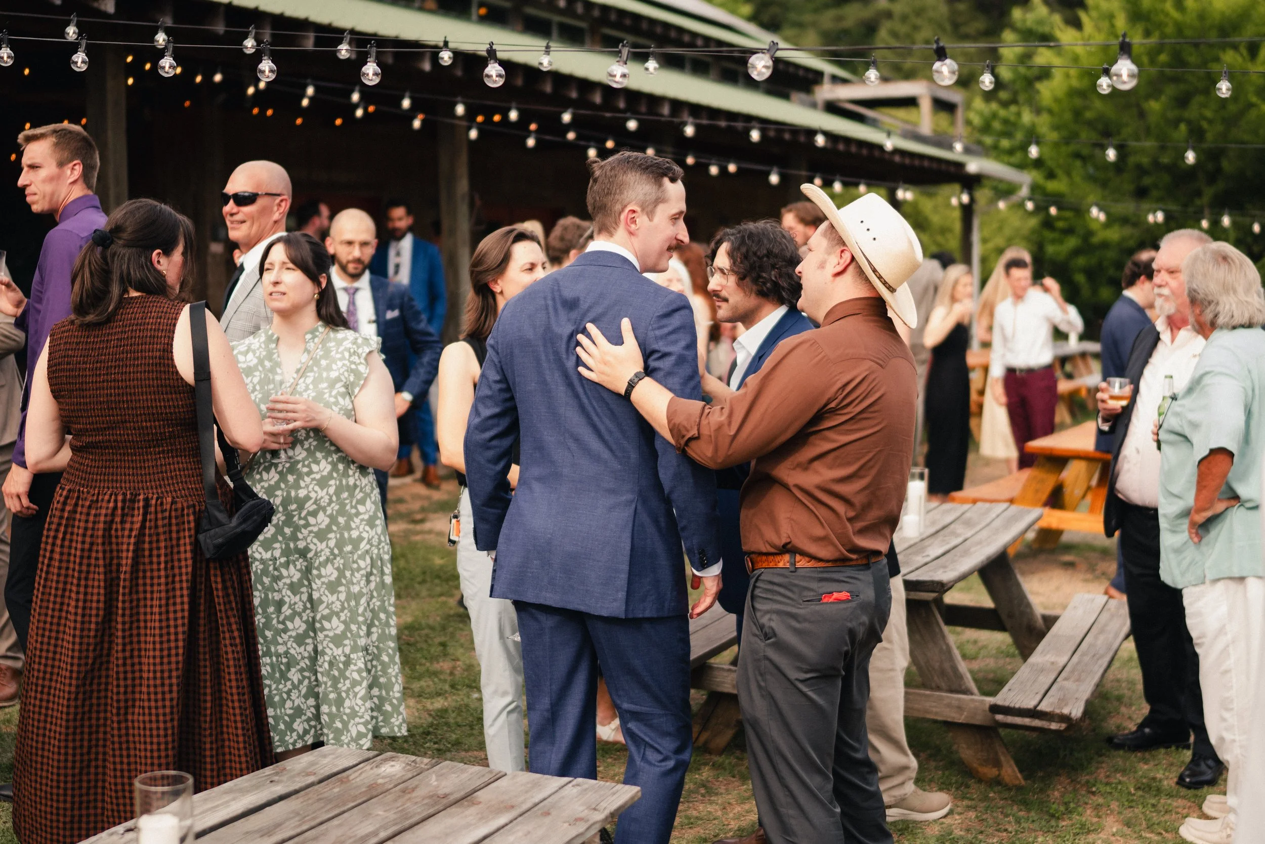 A group of people socializing outdoors at a gathering or party, with string lights overhead, in a setting with trees and wooden picnic tables.