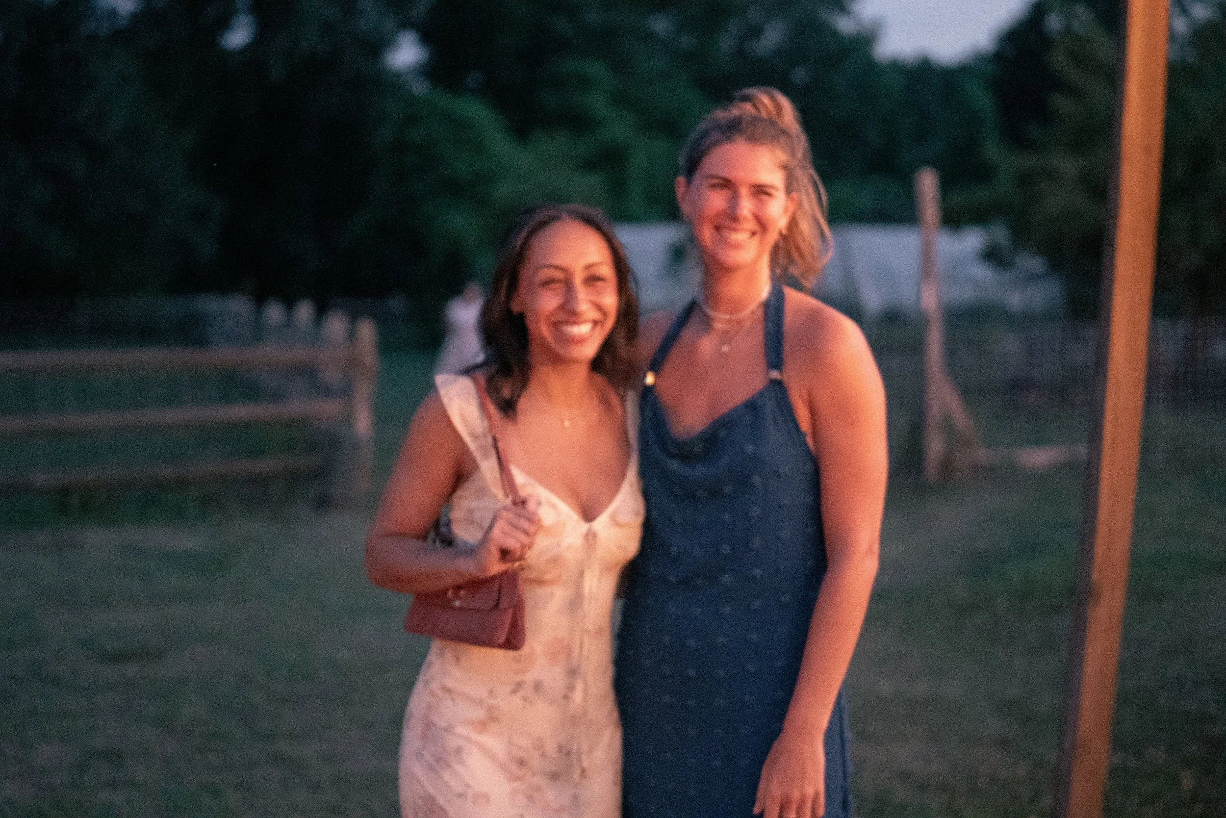 Two women smiling outdoors during evening, one wearing a light dress with spaghetti straps and holding a drink, the other in a blue dress with a halter top.