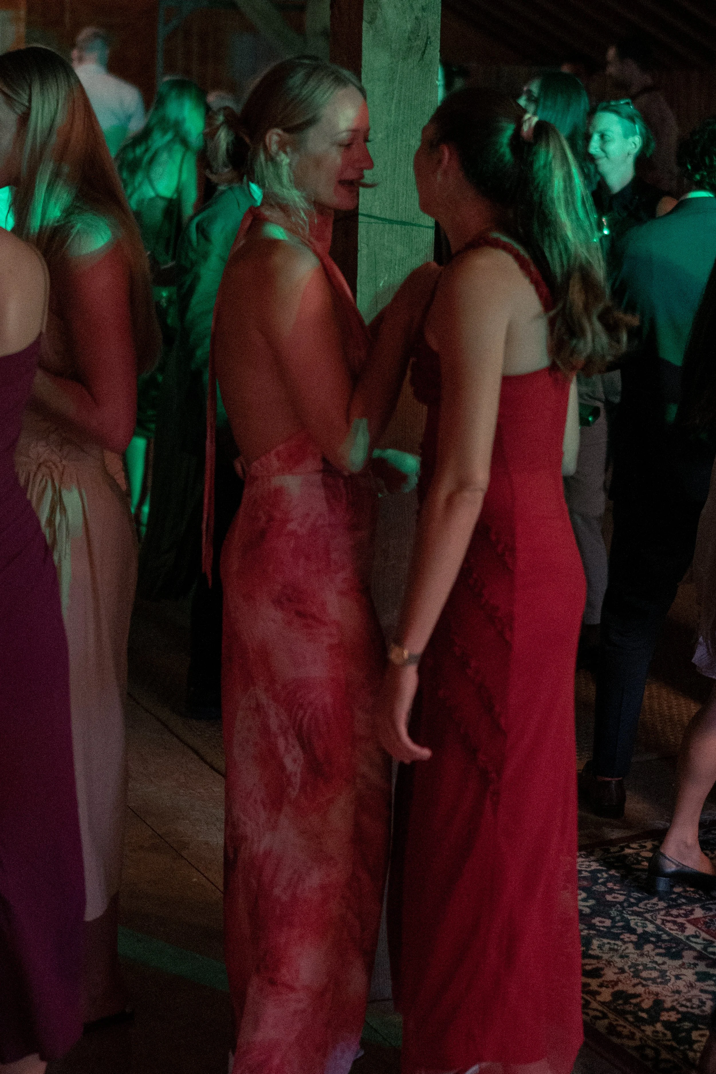 Two women dressed in red lace dresses are standing close, facing each other and smiling at a social gathering or party. Other people are in the background, some dancing or talking, in a dimly lit room.