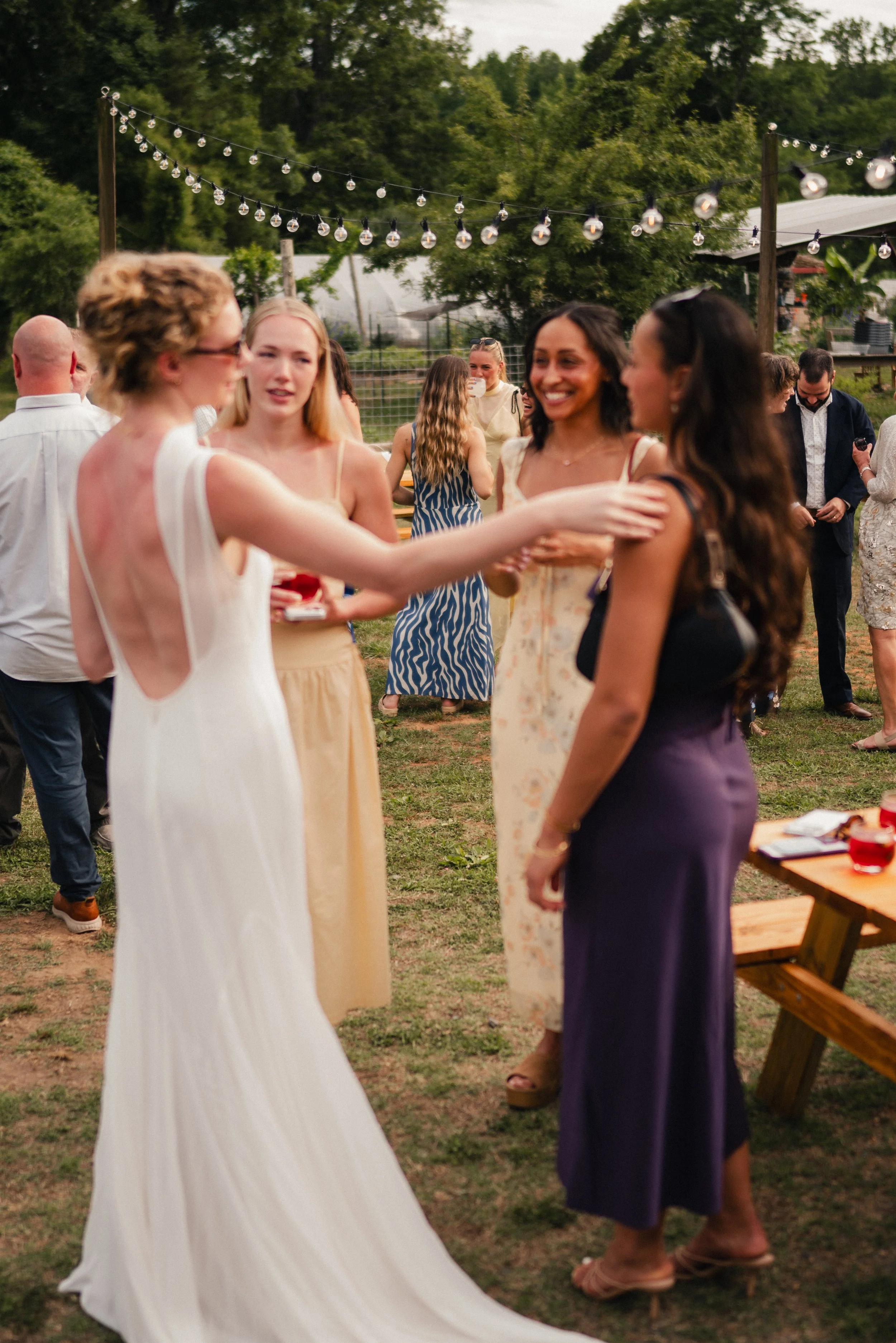 Four women are having a conversation outdoors at a social gathering, with string lights overhead and trees in the background.