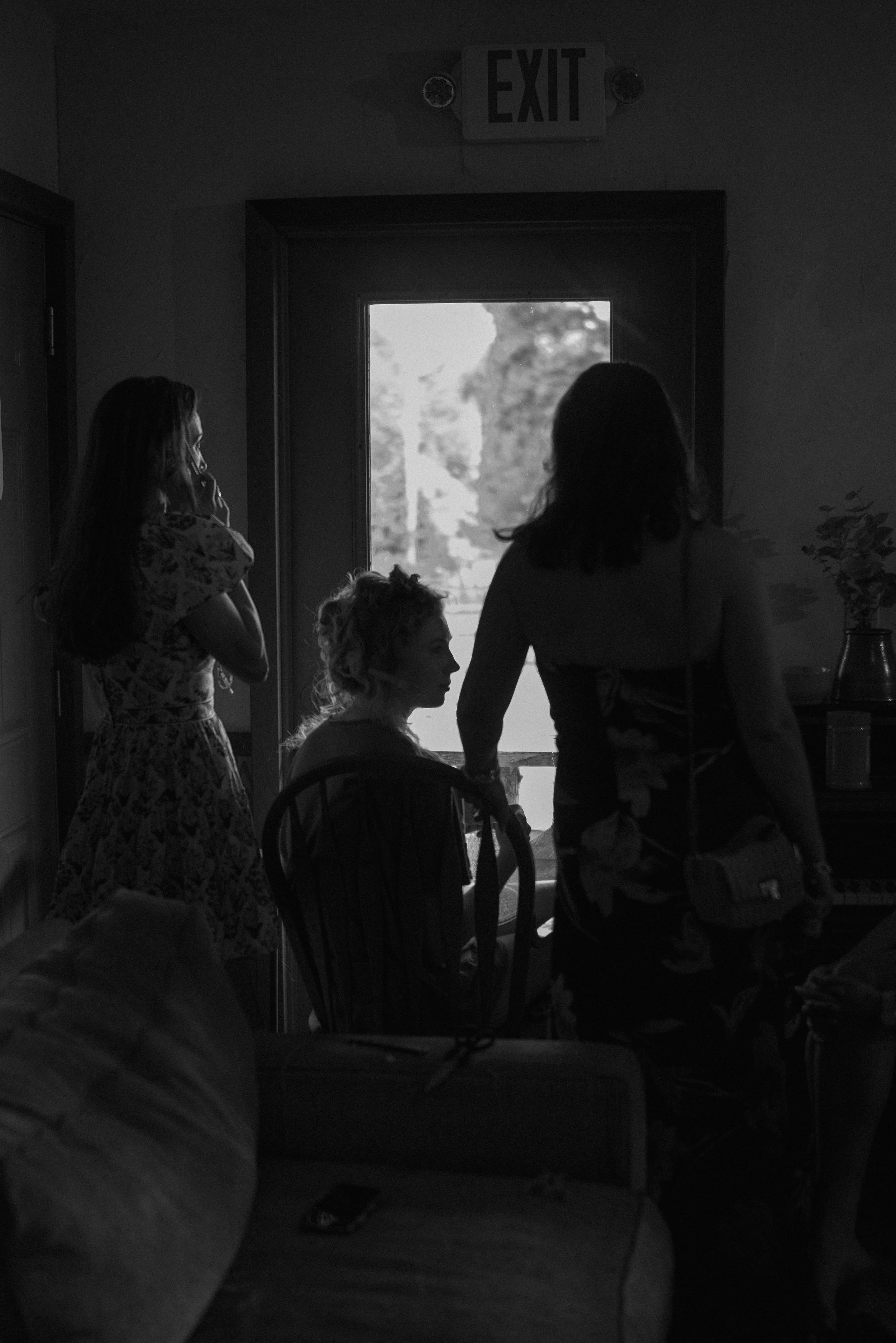 Four people inside a room near an open door, with trees visible outside, and an EXIT sign above the door. One person is standing, three are seated, and one person is wearing a backpack.