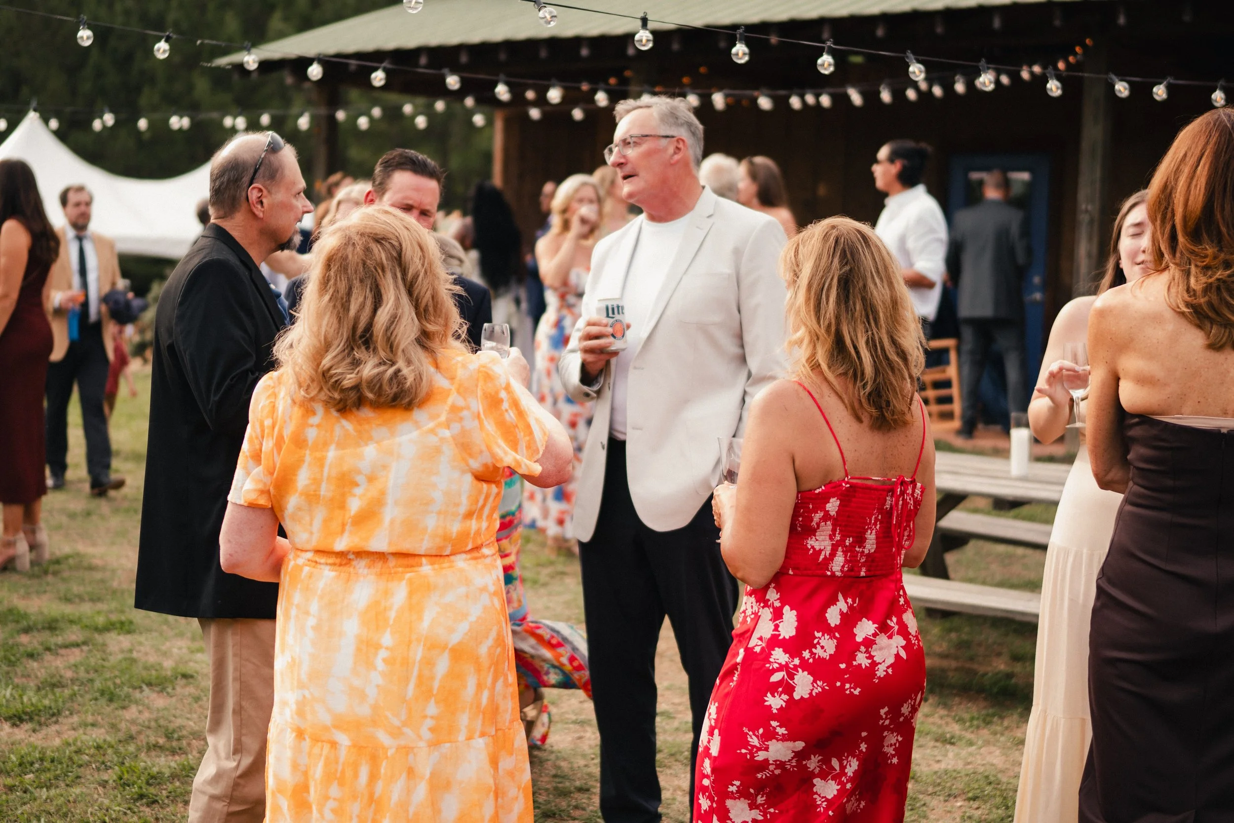 Group of people at an outdoor social gathering, dressed in summer attire, standing and talking under string lights with a wooden structure in the background.