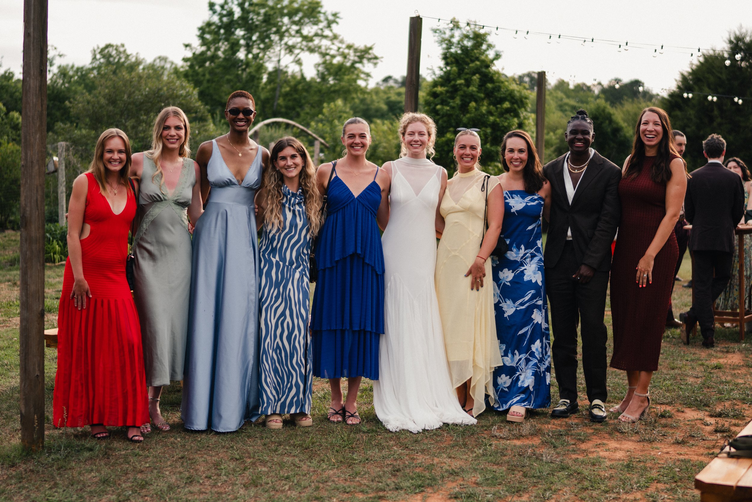 Group of women dressed in formal and summery dresses standing outdoors, smiling for a photo, with trees and string lights in the background.