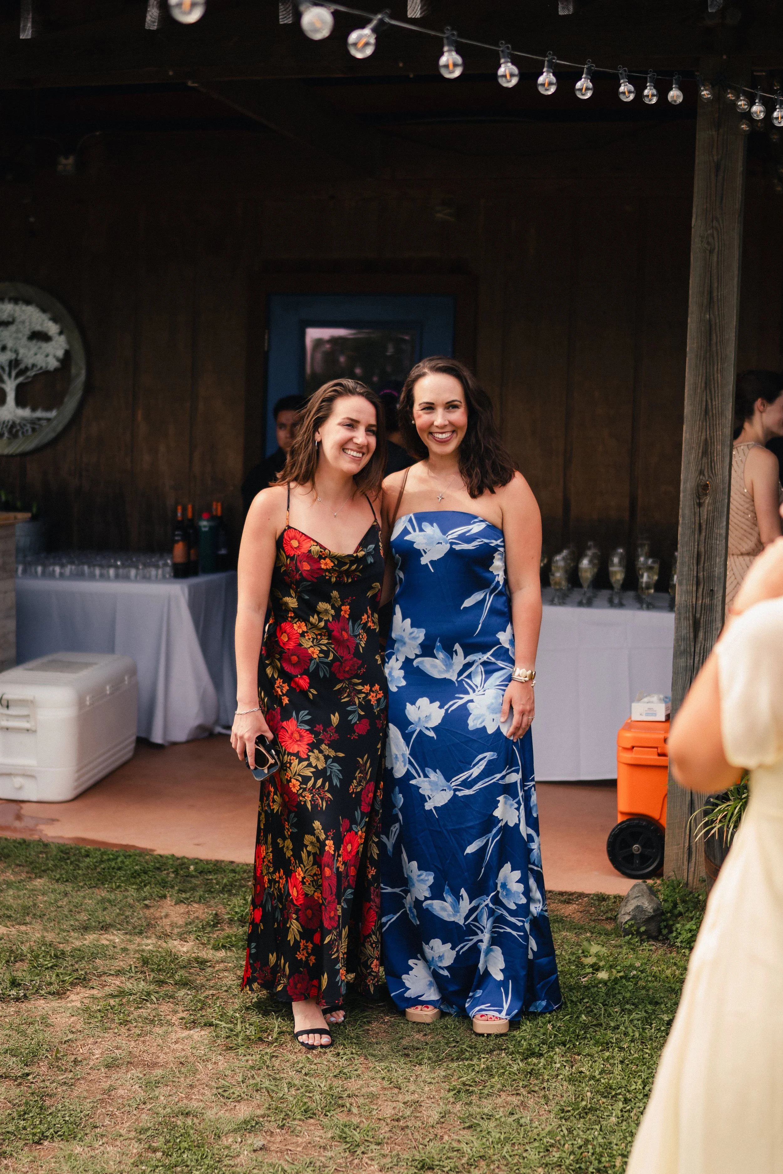 Two women in floral dresses standing together outdoors at a celebration, smiling for the camera.
