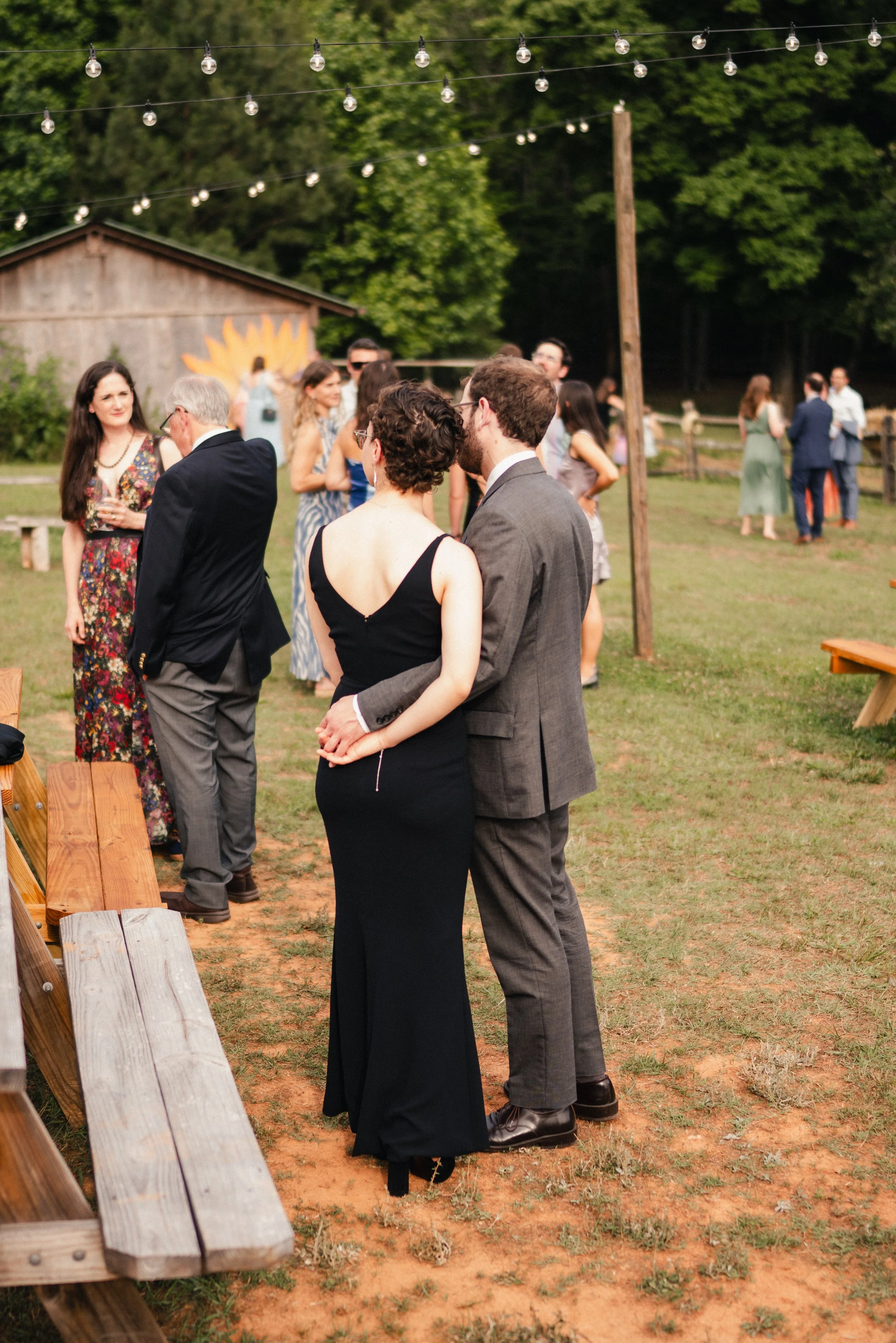 People socializing at an outdoor event under string lights with a rustic building and green trees in the background.