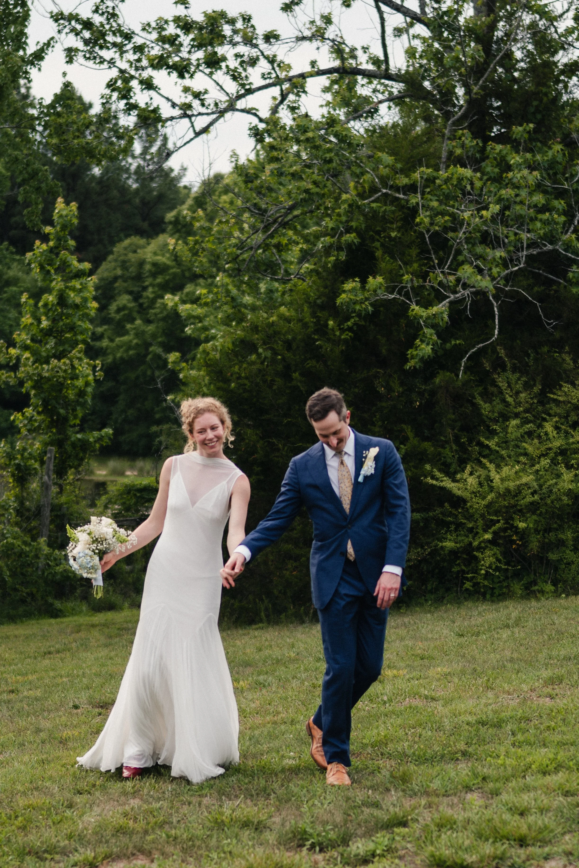 A bride and groom holding hands, walking on grass outdoors during a wedding celebration, with a backdrop of green trees and overcast sky