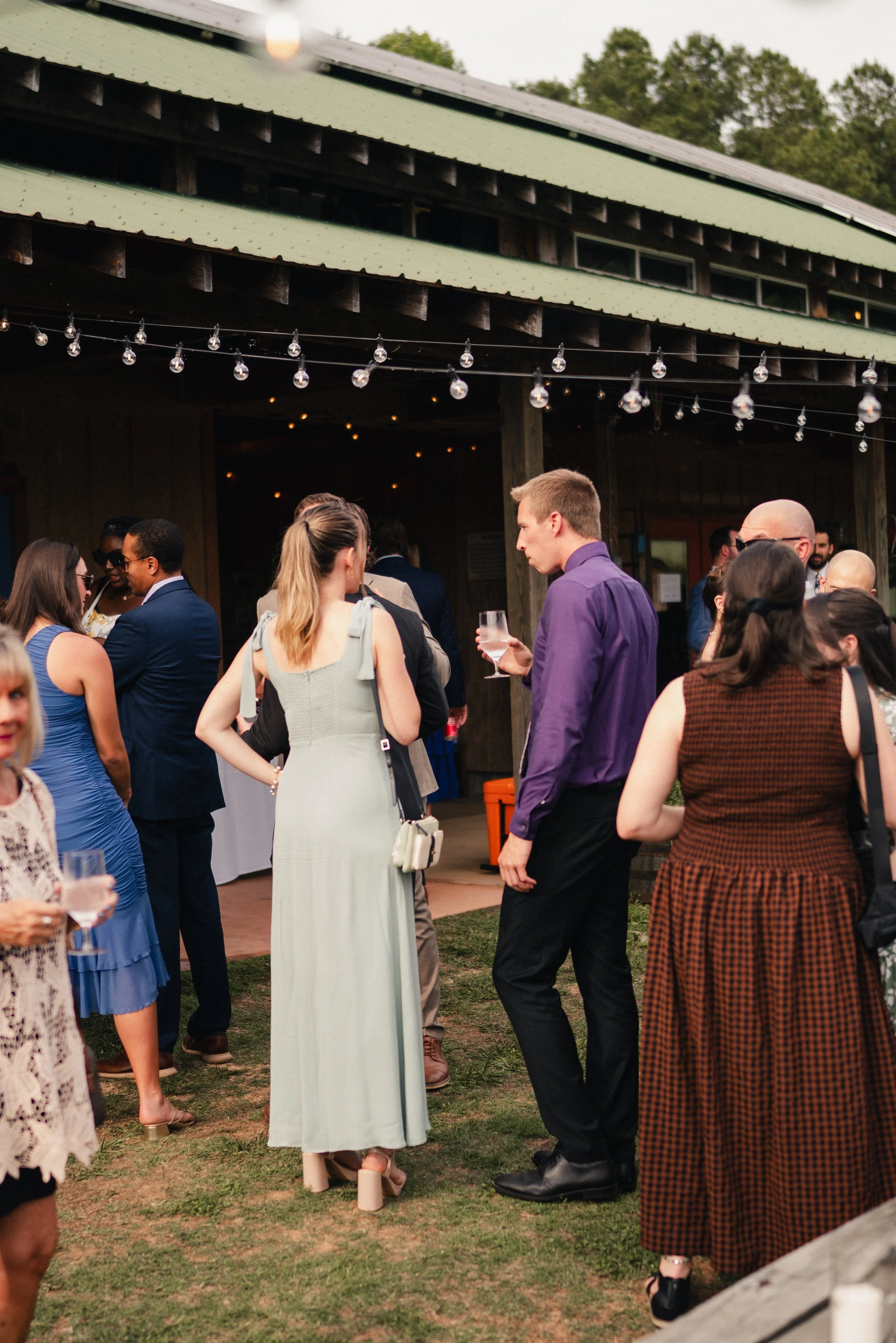 People gathered outdoors at a social event or wedding reception, talking and holding drinks under string lights near a barn or rustic building.
