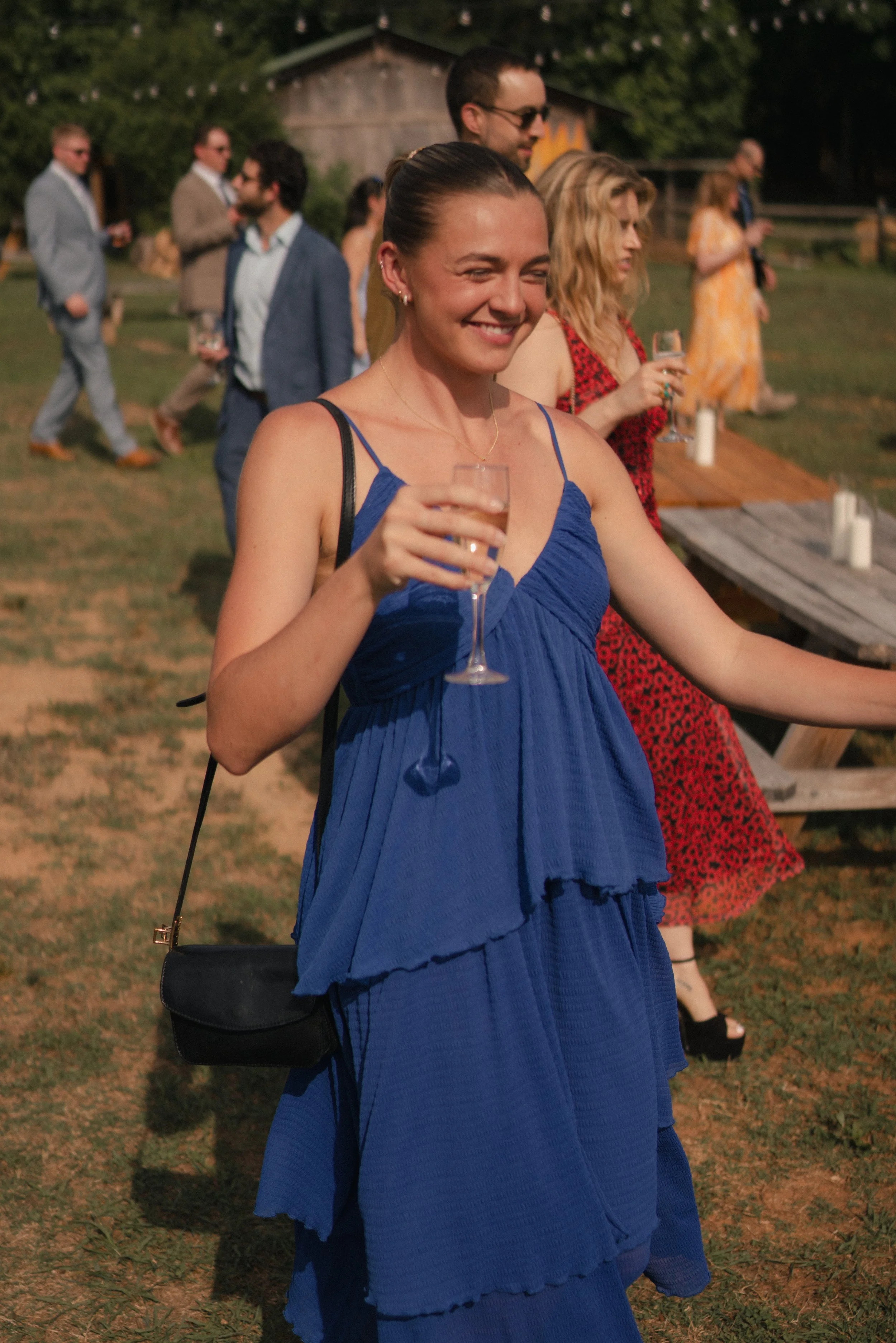 A woman smiling and holding a glass of champagne at an outdoor gathering during sunset. She is wearing a blue dress and has a black purse with a gold clasp. In the background, several people are socializing, and there is a rustic barn with trees arou