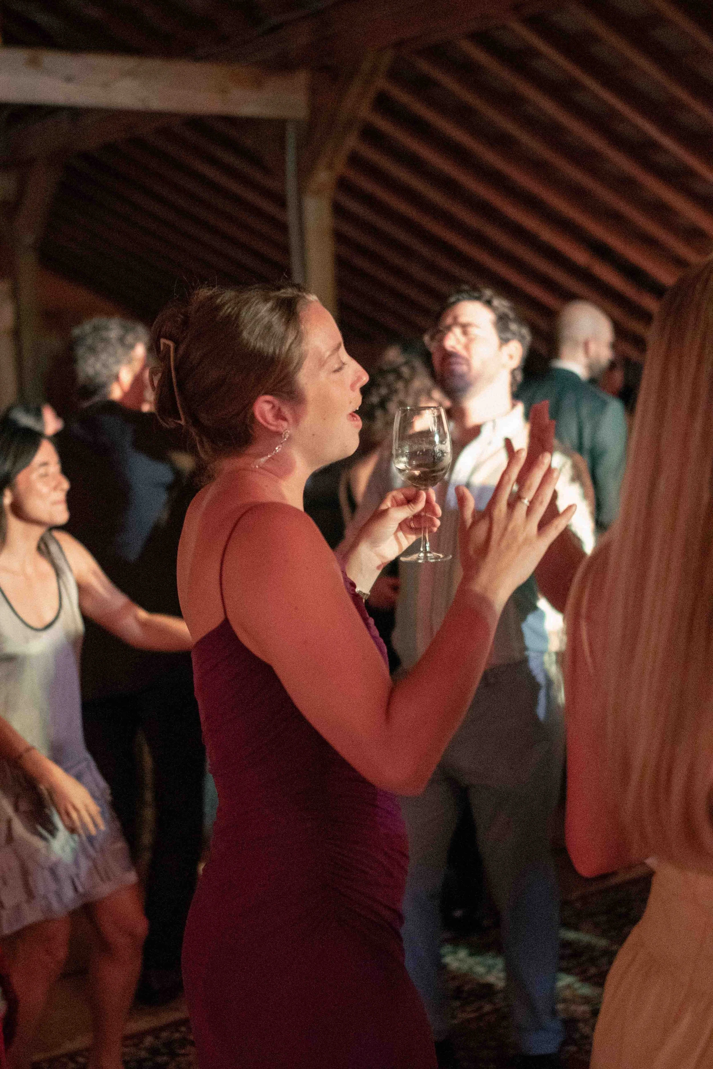 A woman in a red dress holding a glass of wine, dancing at a social gathering with other people in a wooden-paneled room.