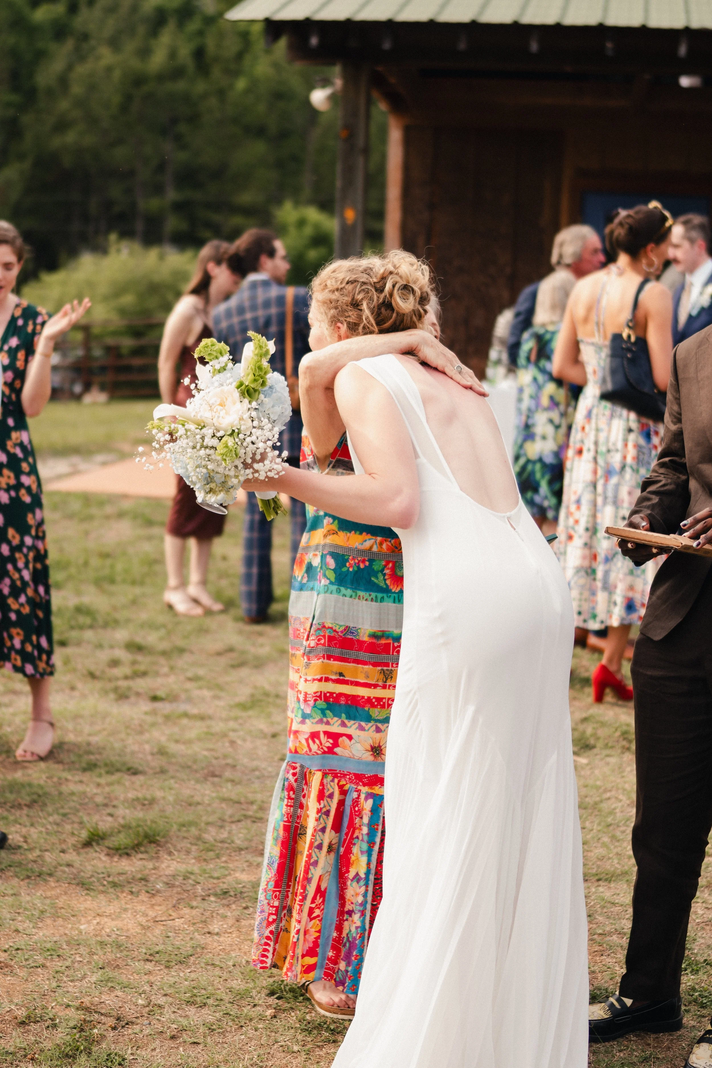 A wedding celebration outdoors with people dancing and talking, a woman in a white dress hugging another woman in a colorful dress, holding a bouquet of flowers, with a group of guests in the background, and a rustic wooden building.