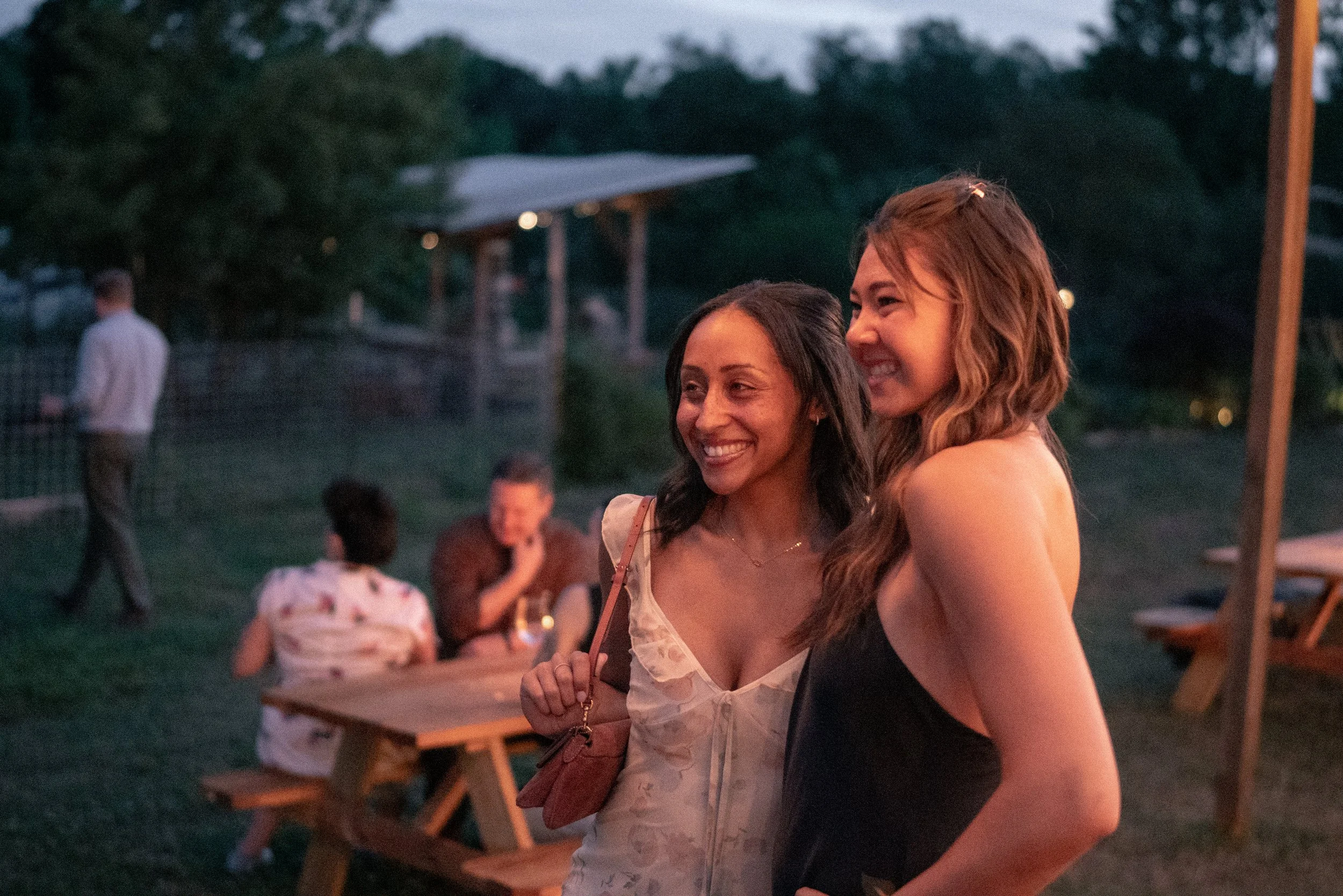 Two women smiling and hugging at an outdoor gathering during evening, with other people and tables in the background.