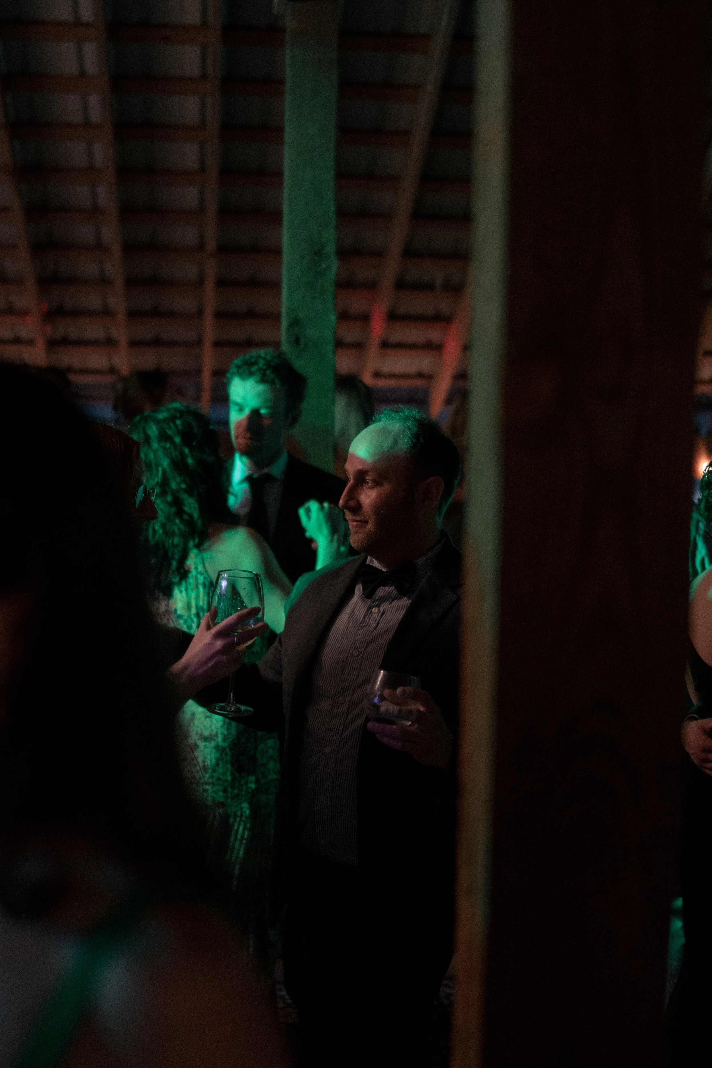 People dressed in formal attire, holding glasses of drinks, at a dimly lit indoor event or party, with a wooden ceiling structure visible in the background.