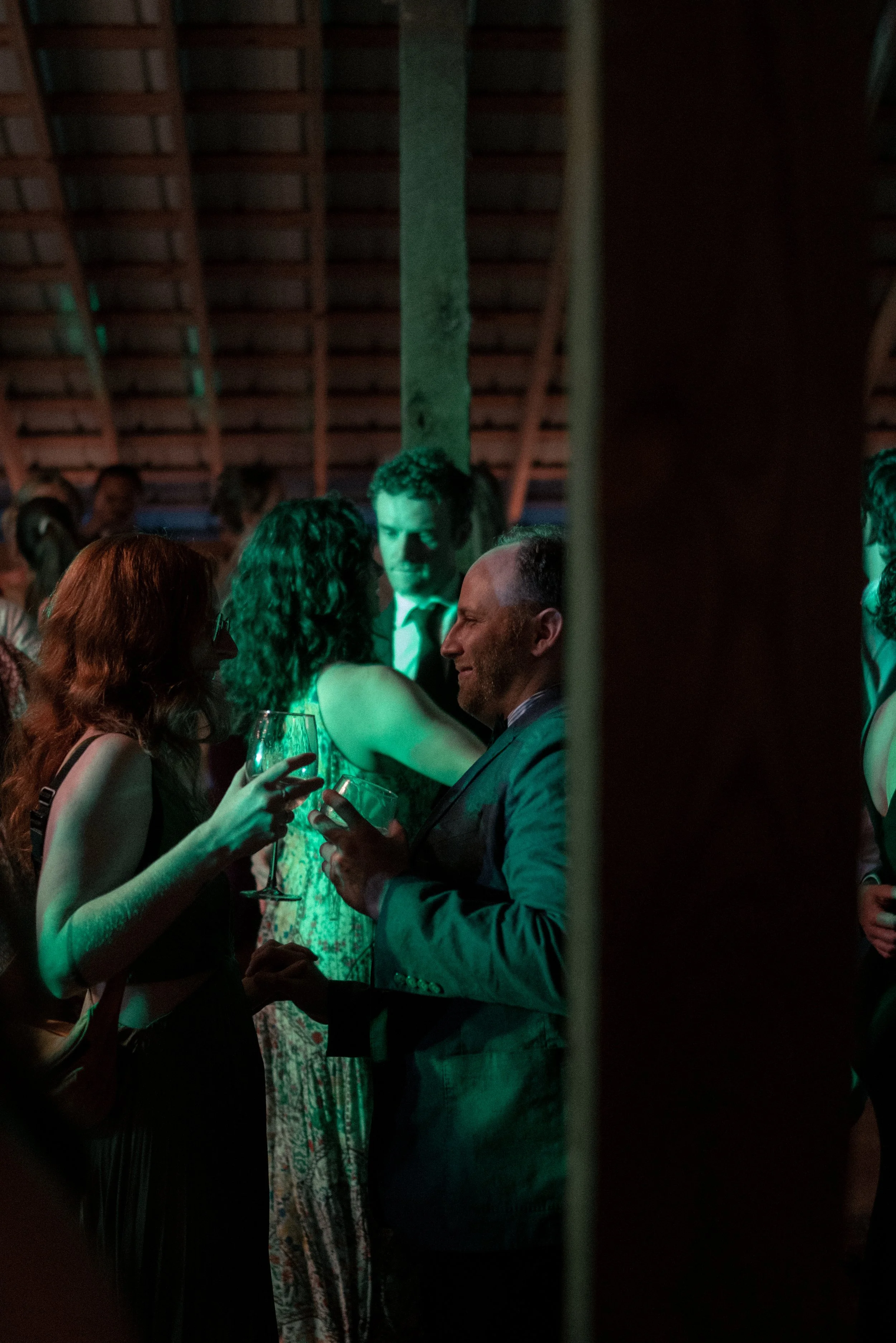 A group of people socializing at an indoor event, illuminated with green lighting, with a view of the ceiling with wooden beams. Two women are holding wine glasses and talking, while a man in a suit is smiling and engaging conversation.