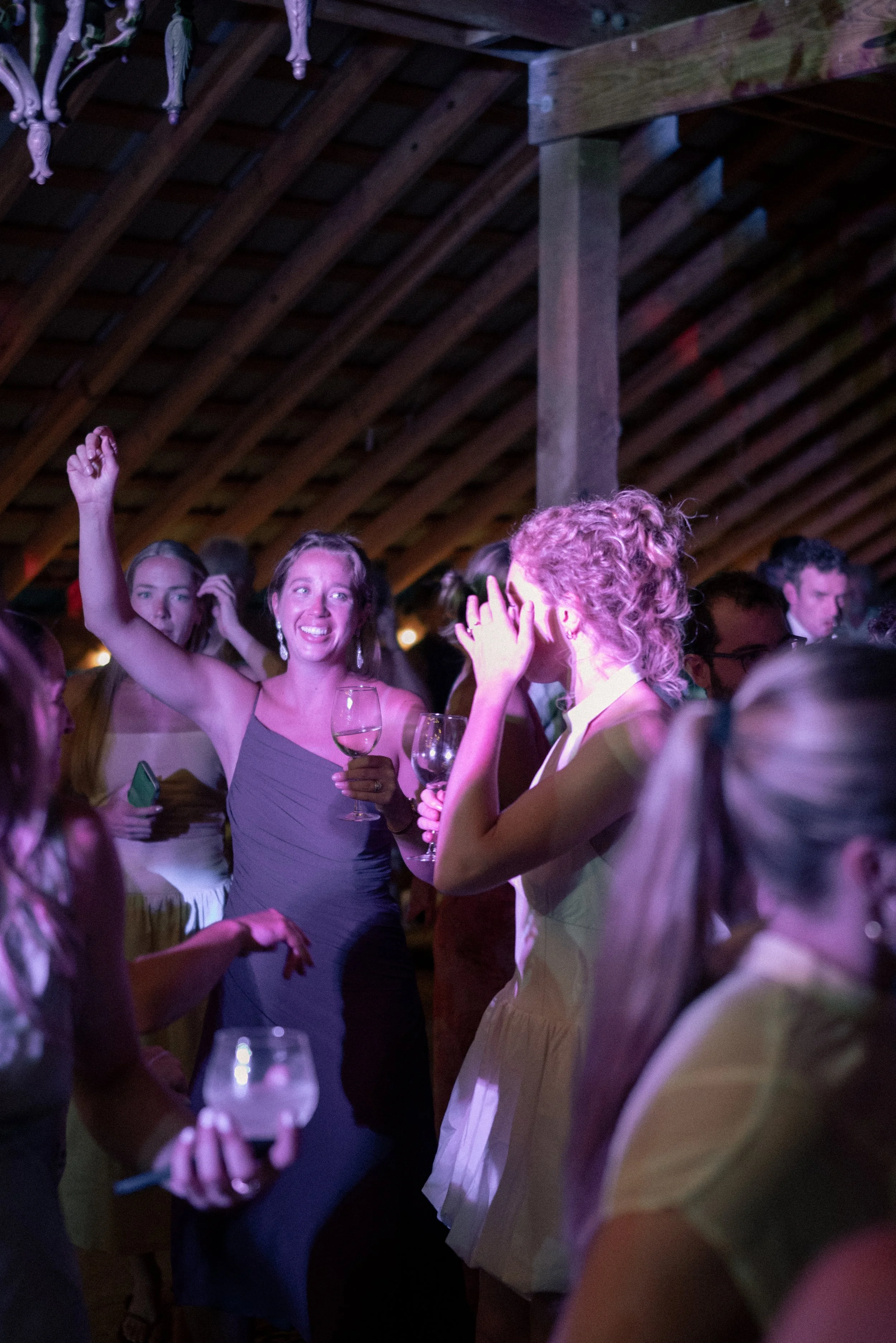 People socializing at a party or wedding reception, some holding glasses of wine, in a dimly lit venue with a wooden ceiling, smiling and engaging in conversation.