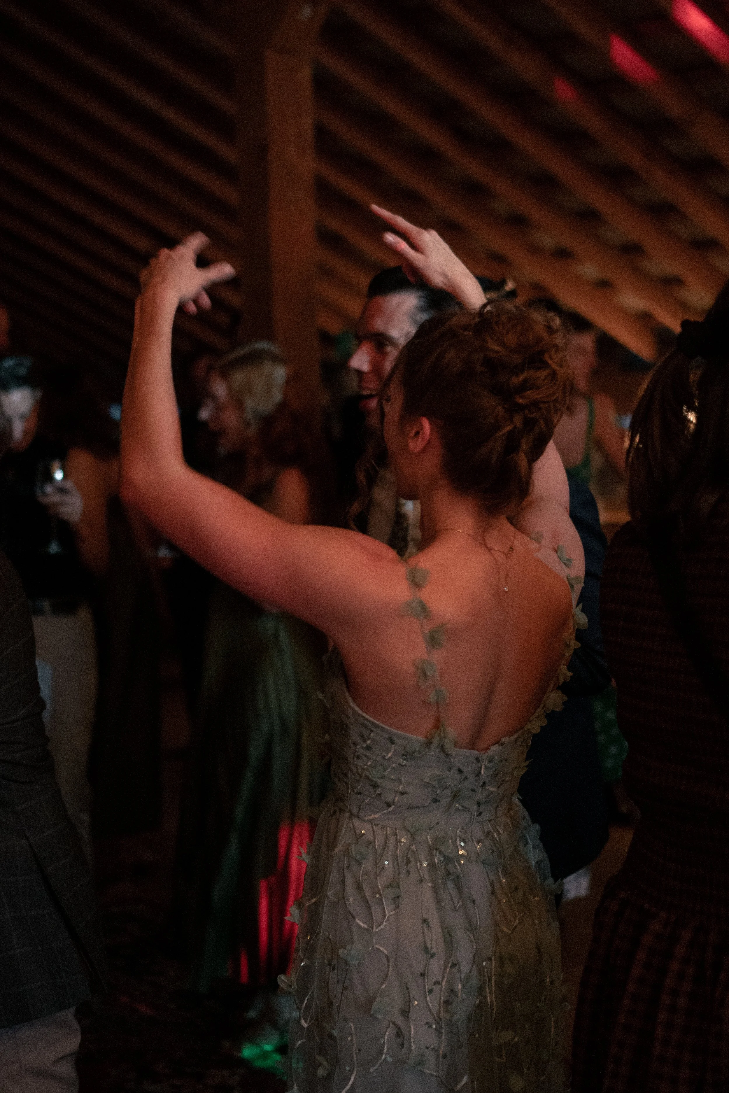 A woman with auburn hair styled in an elaborate updo dancing at a party, wearing a dress with floral embroidery and sheer fabric, surrounded by other guests in a dimly lit setting.