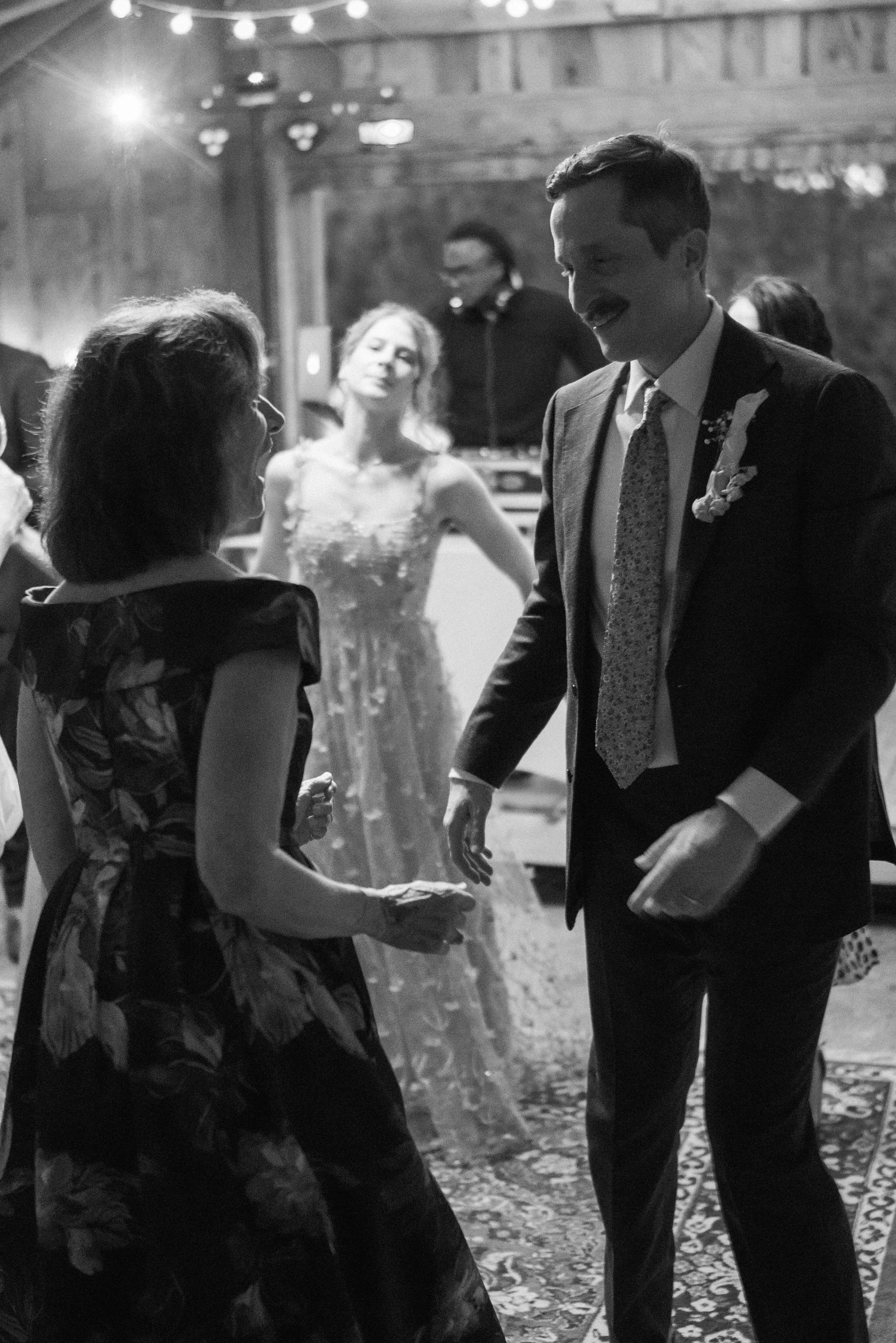 A black and white photo of a wedding reception, showing a man in a suit and a woman in a gown talking and smiling. Other guests are visible in the background, and the setting has string lights and a decorated floor.