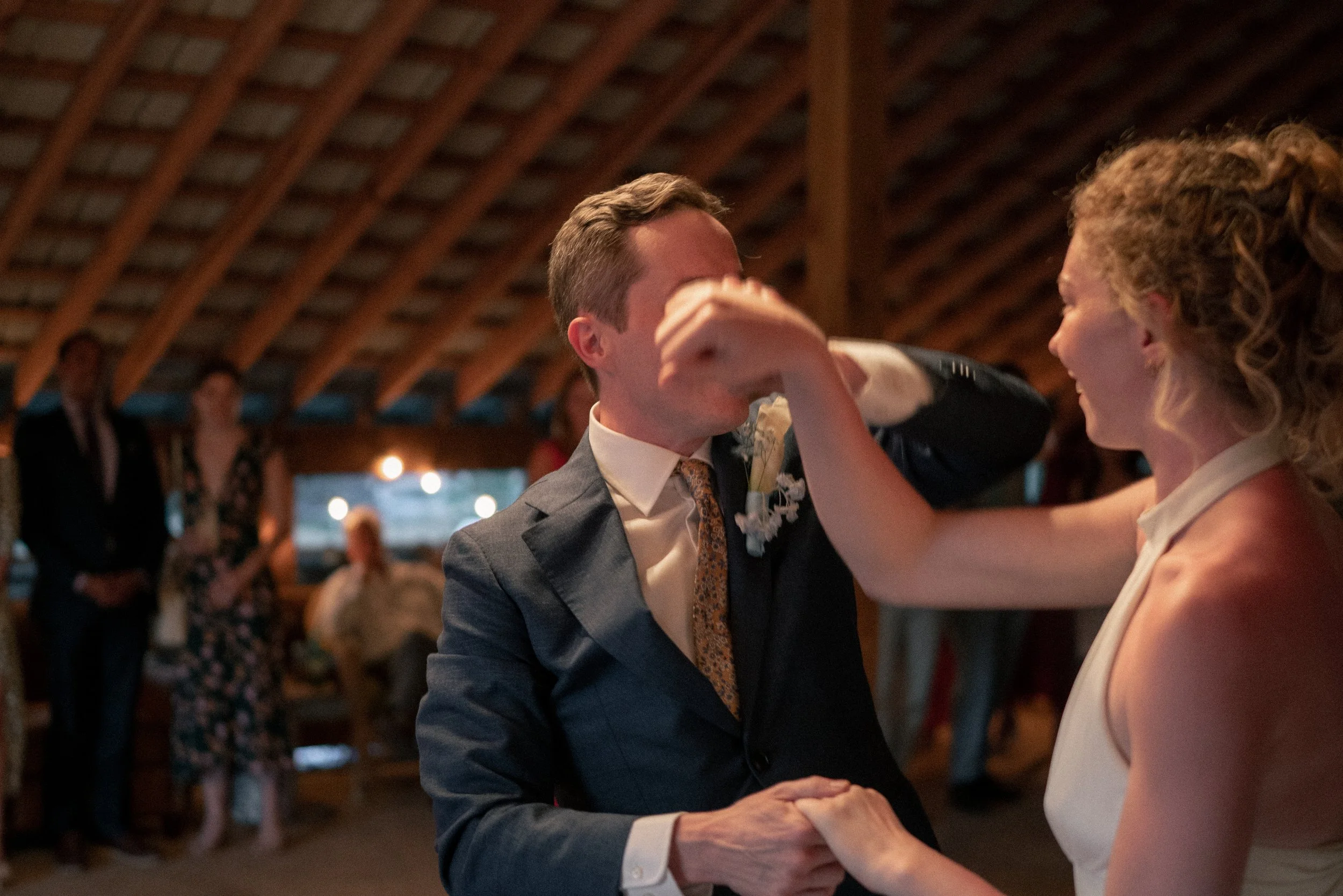 A couple sharing a dance at a wedding reception in a rustic venue with a wooden ceiling. The groom in a suit and tie is dancing closely with a woman in a white dress, surrounded by other guests.