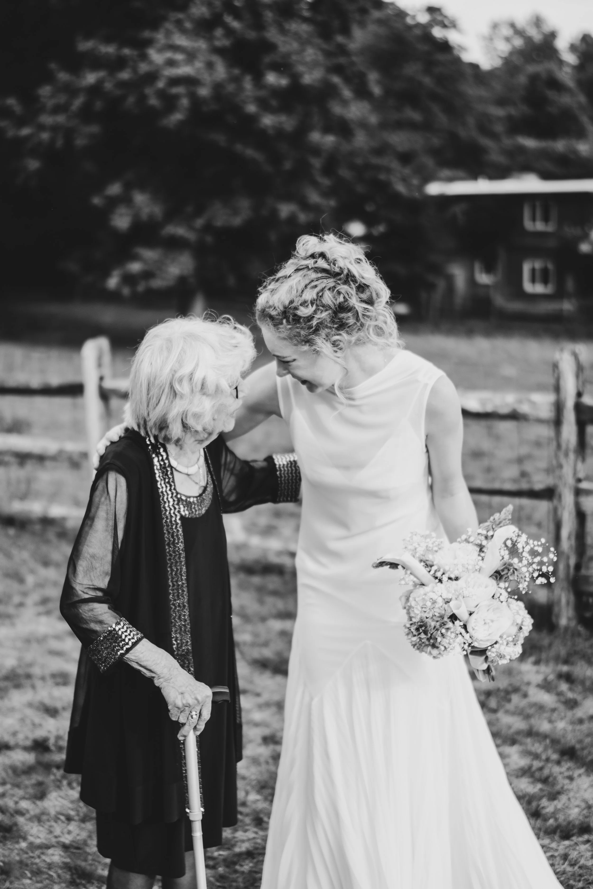 A young woman in a white wedding dress smiling and leaning towards an elderly woman who is in a black dress, also smiling and holding a cane, outdoors in a park-like setting with a house and trees in the background.