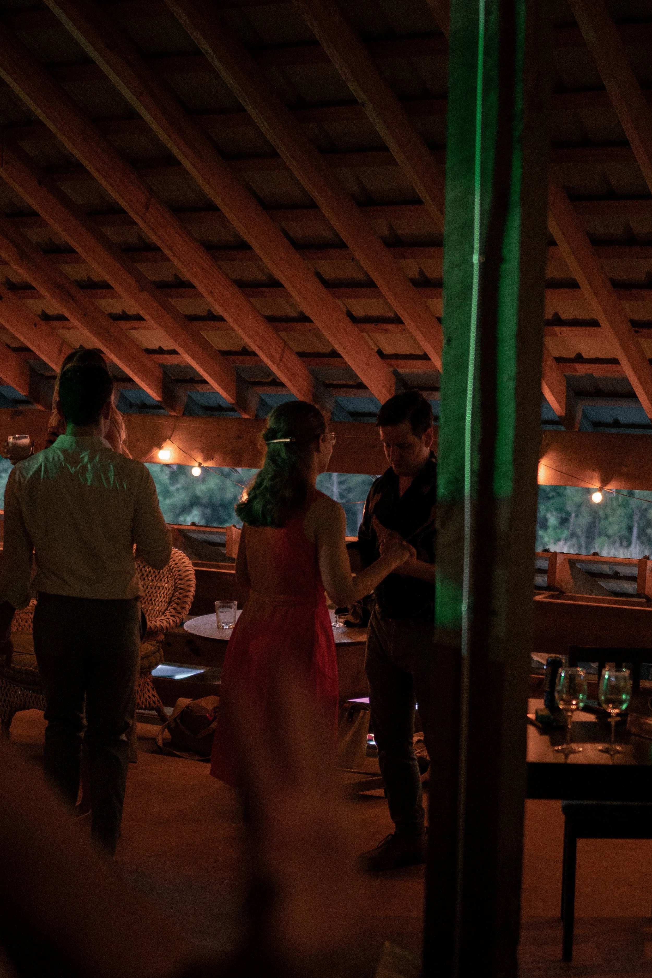 People socializing at a dimly lit indoor party with string lights, a wooden ceiling, and a bar counter on the right side.