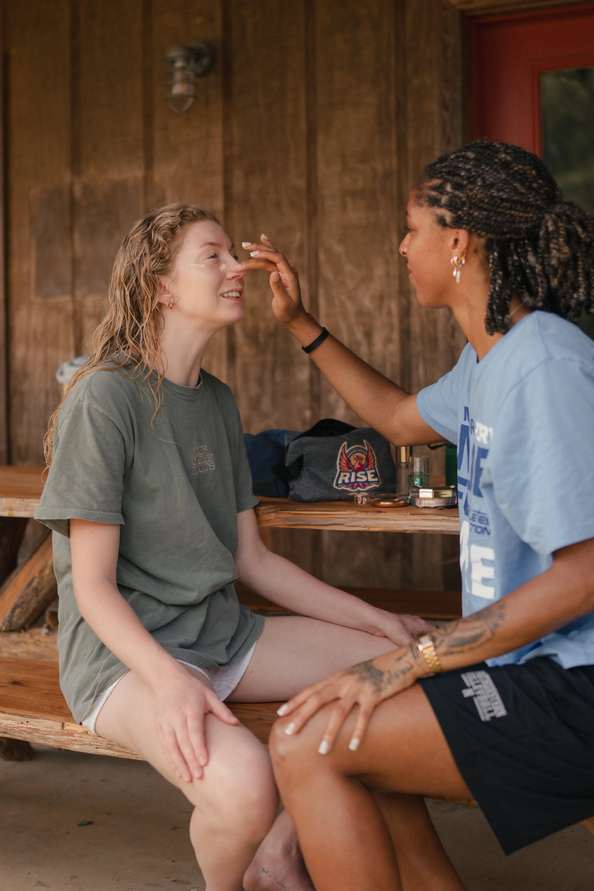 Two women are sitting at a wooden table on a porch with wooden walls. One woman with curly red hair is smiling while another woman with braided hair is touching her nose. The second woman has tattoos on her arms and is wearing earrings.